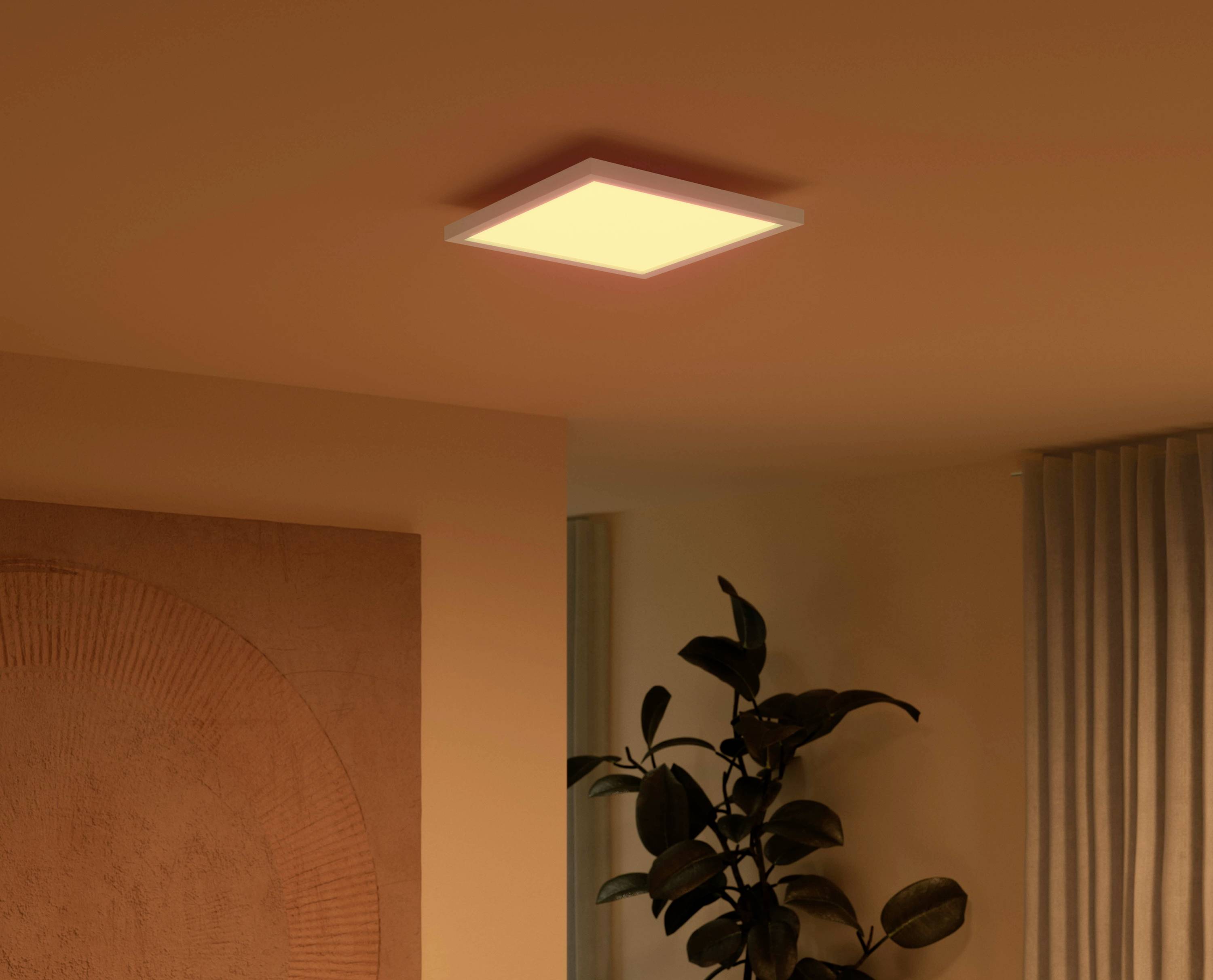 A square ceiling light glows warm white in a cosy room with a plant and curtains in the background.
