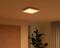 A square ceiling light glows warm white in a cosy room with a plant and curtains in the background.