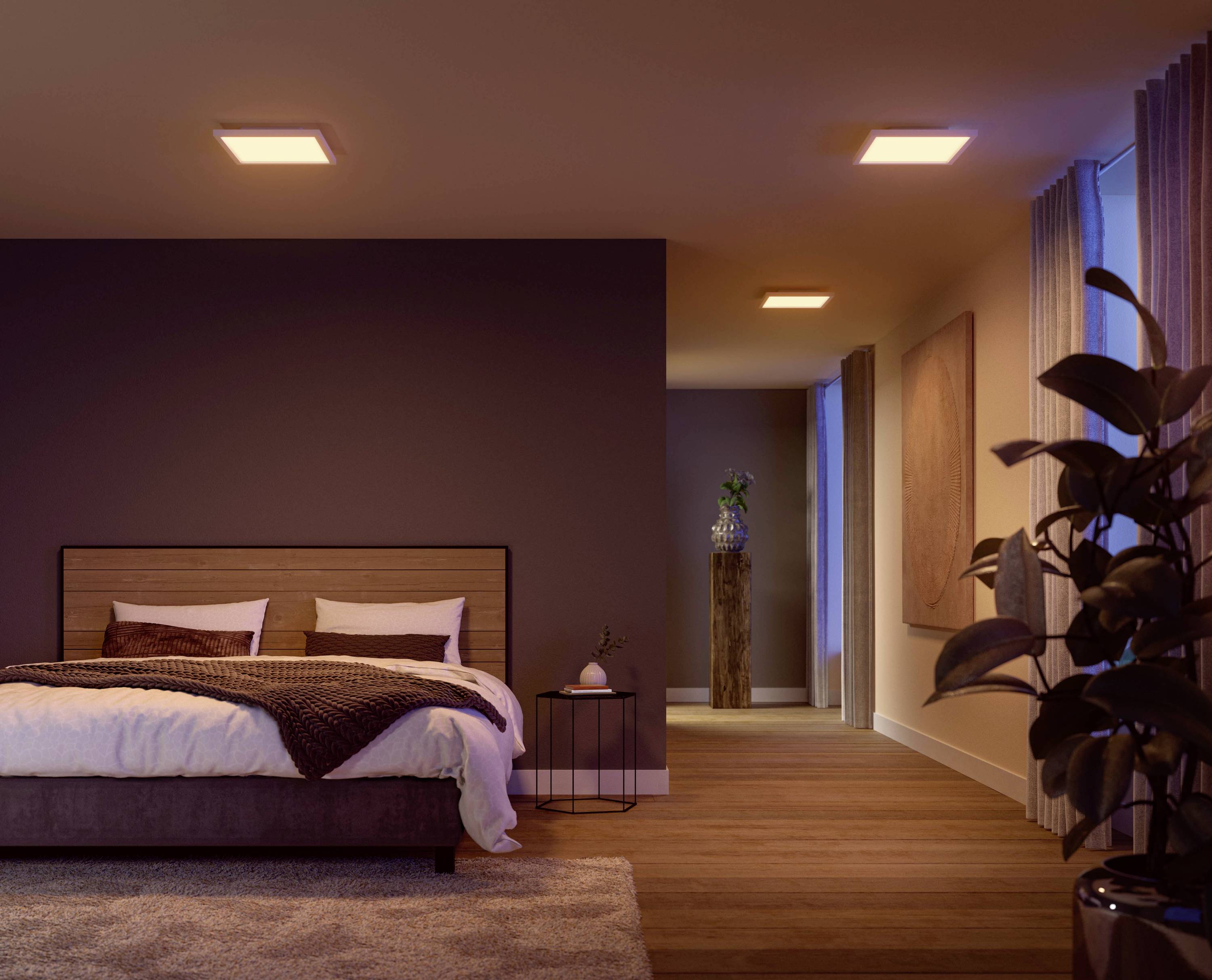 Bedroom with a double bed on the left, cosily lit, fitted carpet, plant on the right, a long corridor leads to an open door at the back.