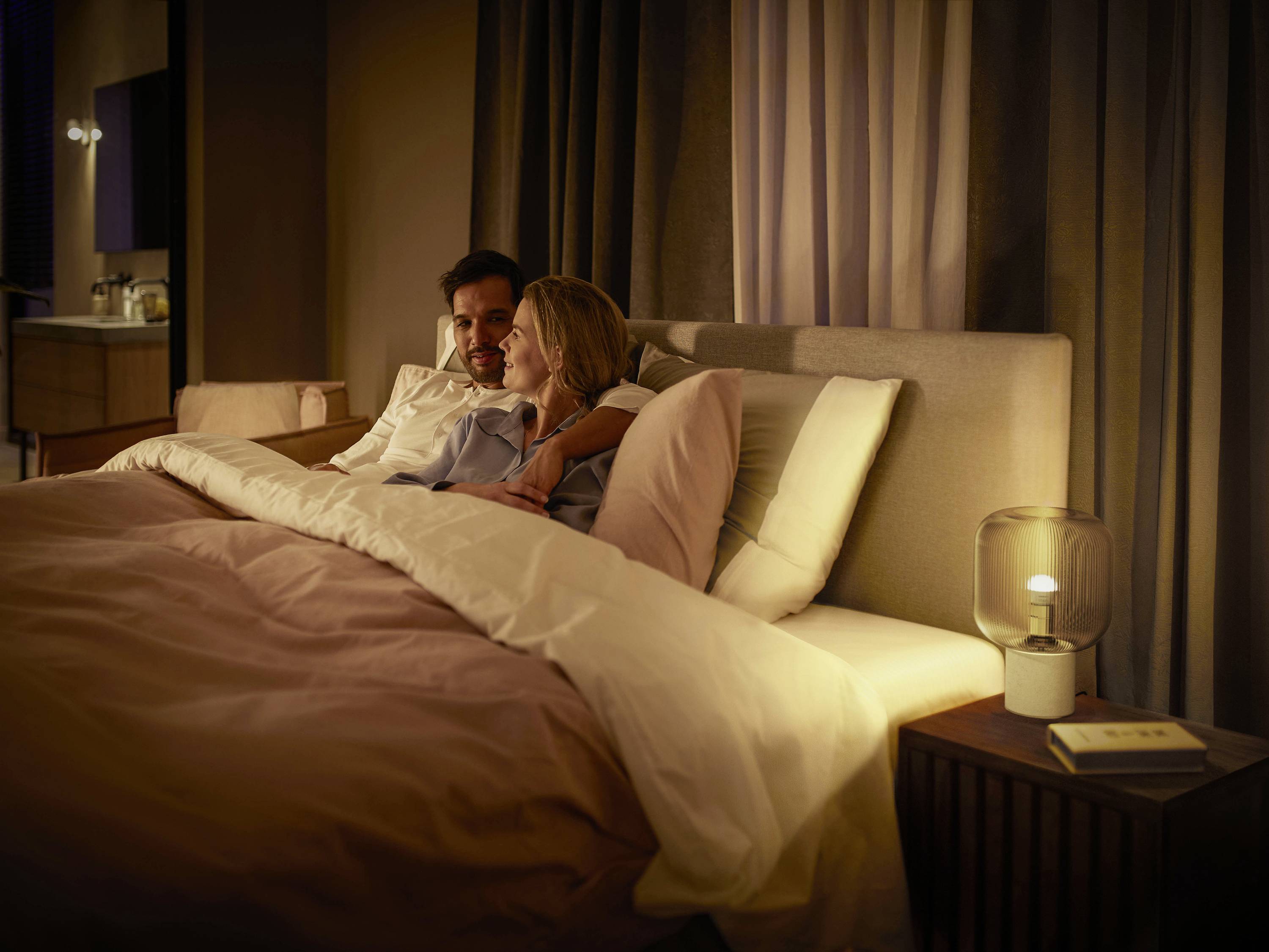 A couple is lying in a cosy bed in a softly lit bedroom. A book and a lamp are placed on the bedside table.