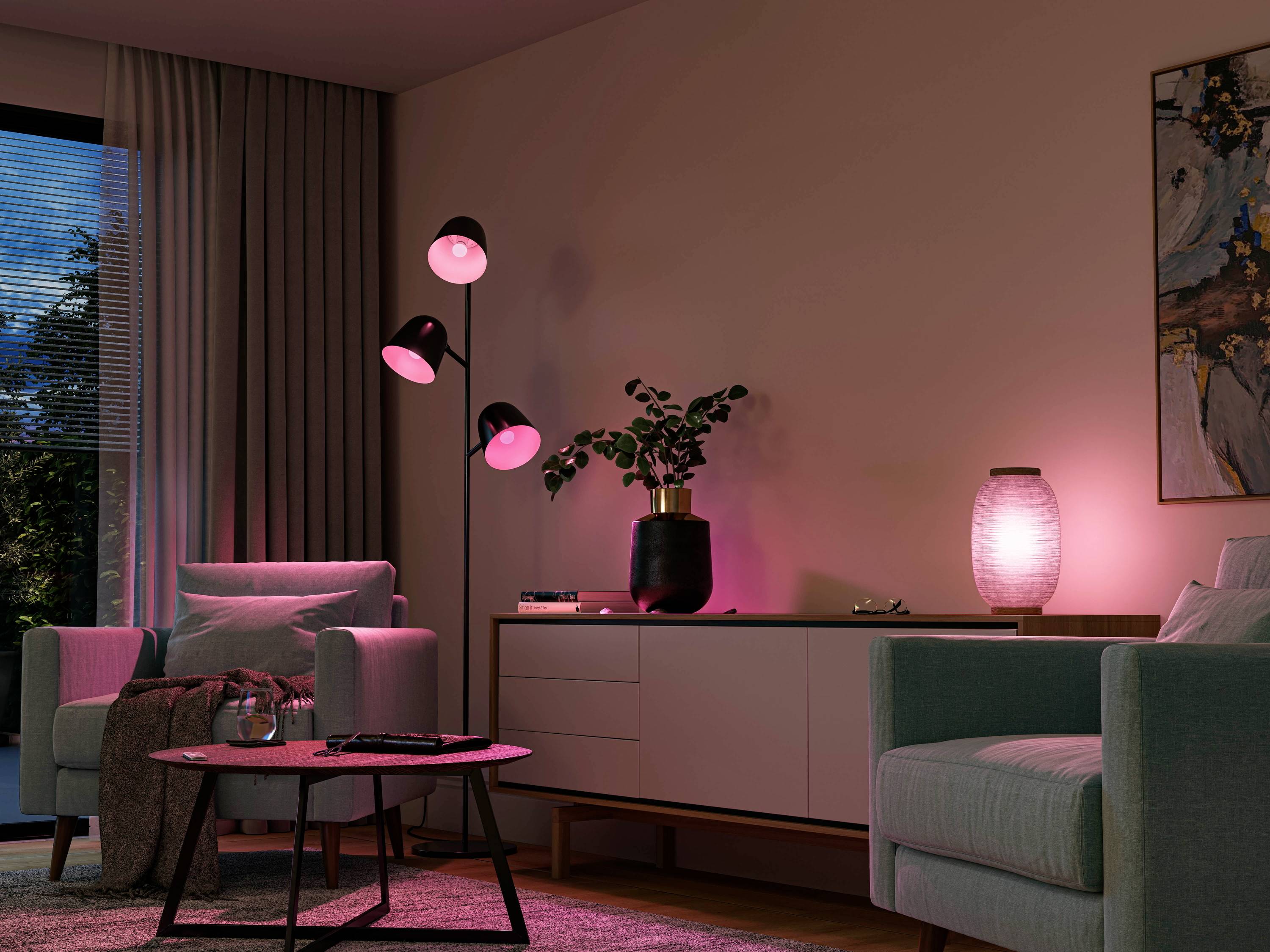 Living room with soft pink lighting, two grey armchairs, round wooden table, standard lamp, chest of drawers with a plant and lamp, modern wall artwork.