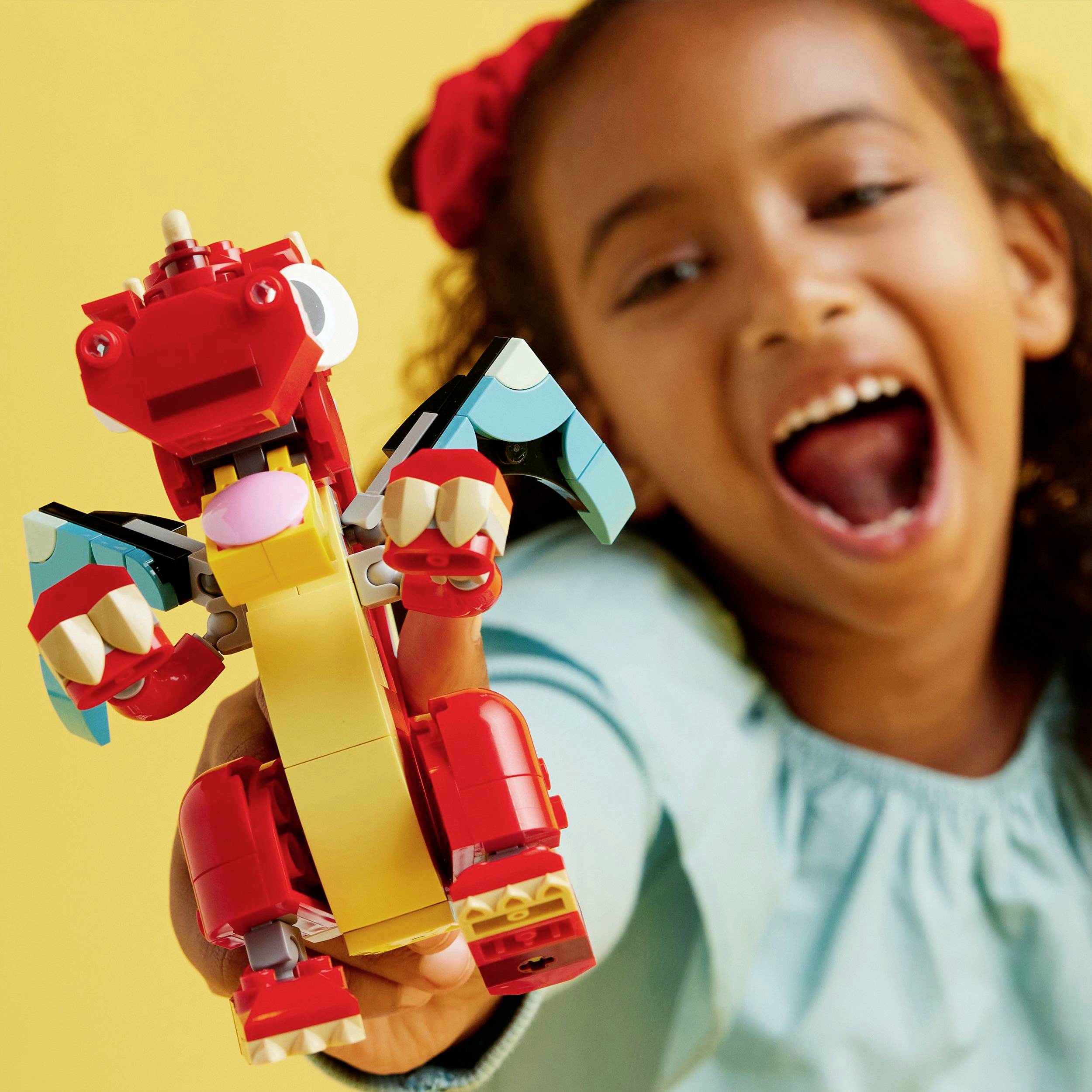 A smiling child is holding a colourful toy dragon made of building blocks. The background is yellow.