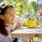 A girl is smiling as she plays with a yellow watering can and toy flowers on a sunny day outdoors.