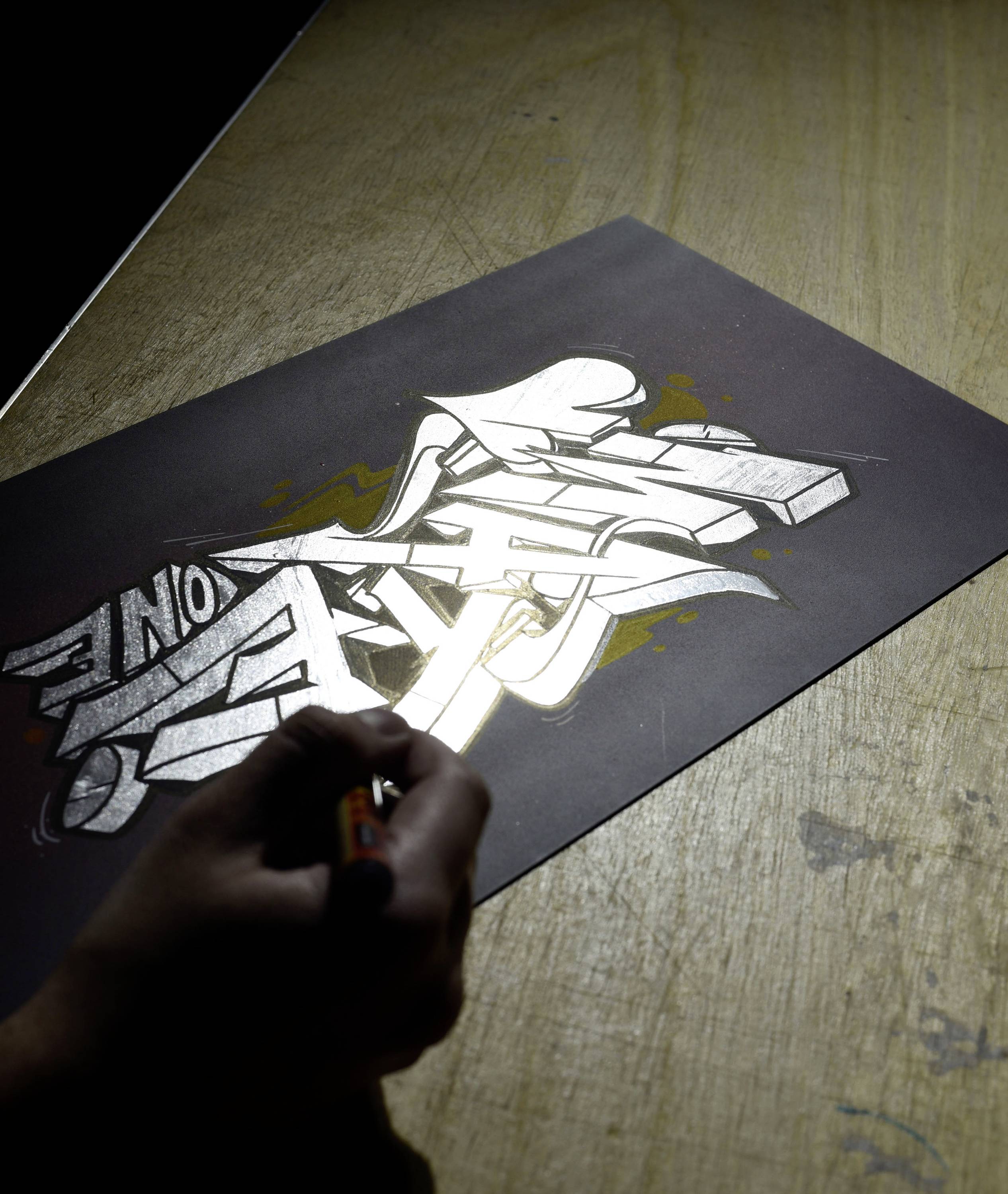 A person is drawing complex graffiti in black and white tones with a pen on a sheet of paper, placed on a wooden table.
