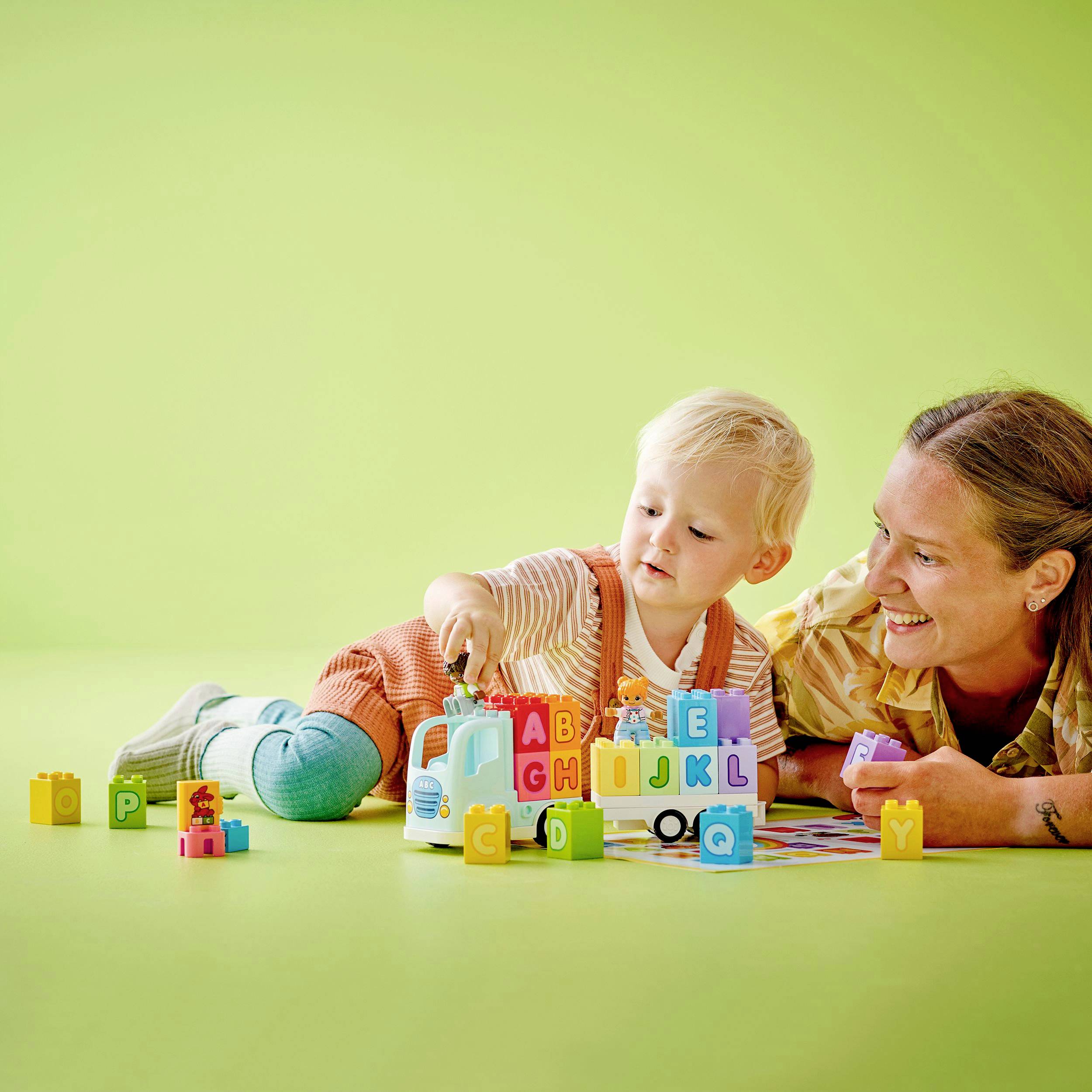 A toddler is playing with alphabet building blocks on a green background, accompanied by a smiling adult who is helping the child.