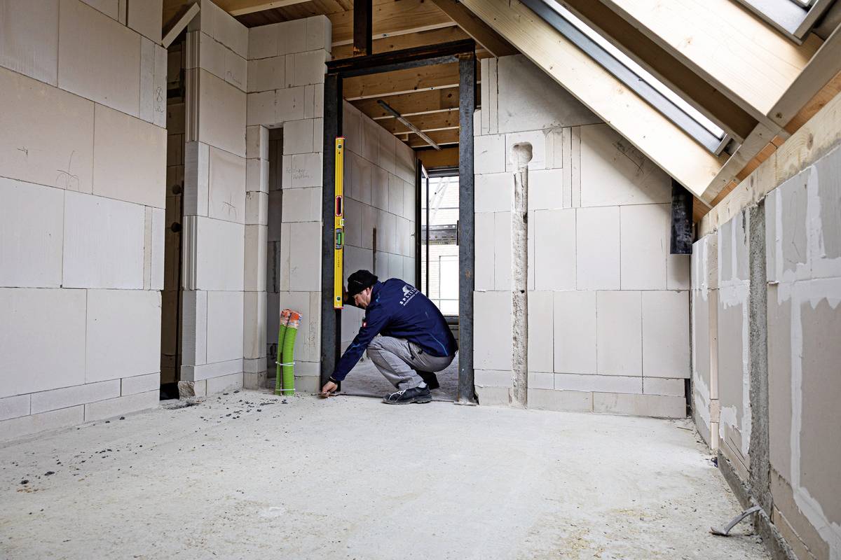 A construction worker checks the floor of a building under construction using a spirit level. Sound insulation and clad walls are visible.