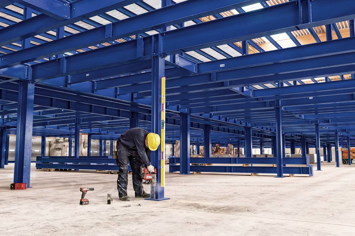 A person wearing a yellow hard hat is working with tools on a blue steel structure inside a large, partially completed building.