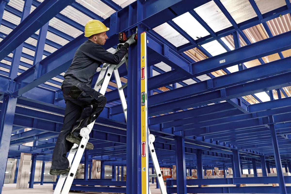 A worker wearing a yellow hard hat stands on a ladder, measuring a blue steel frame inside an industrial hall.