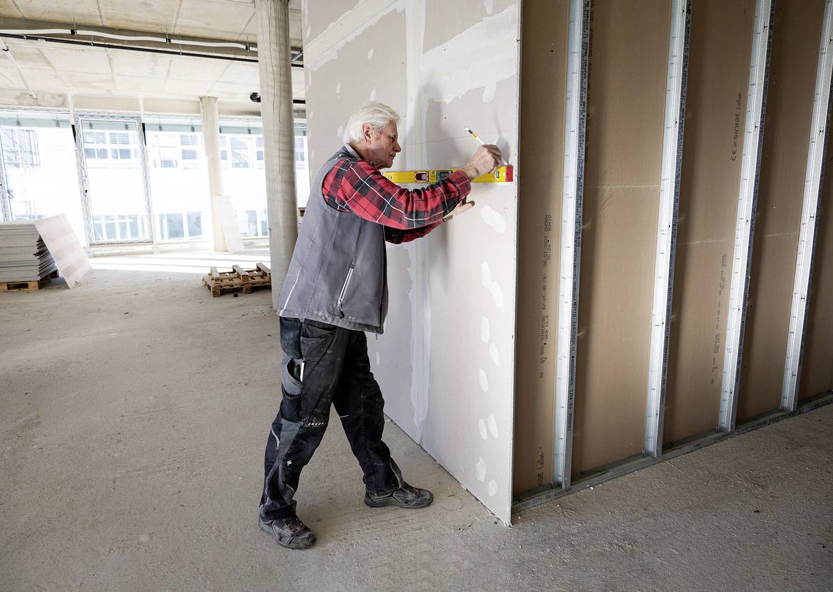 A person is checking the alignment of a plasterboard wall with a spirit level in an unfinished room.