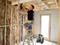 A tradesman wearing protective headphones is standing on a ladder and drilling into the ceiling of a timber-framed structure in a house under construction.