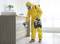 A person in a yellow protective suit disinfects a surface with a spray device in an office. Bright windows are visible in the background.
