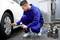A mechanic in blue overalls is checking the tyre pressure of a white car using a portable air compressor.