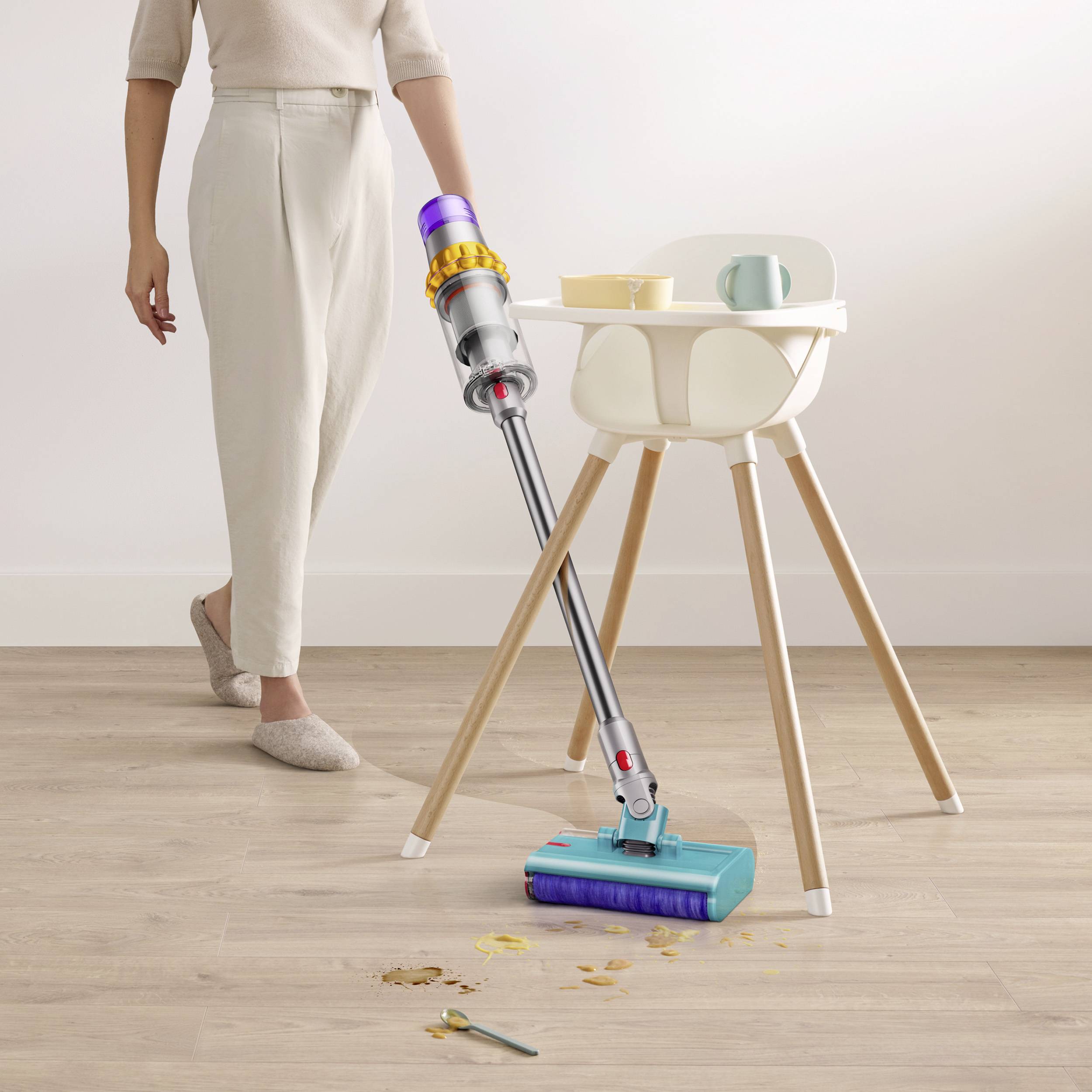 A person is hoovering crumbs around a high chair. The floor is light-coloured and the vacuum cleaner has a colourful head.
