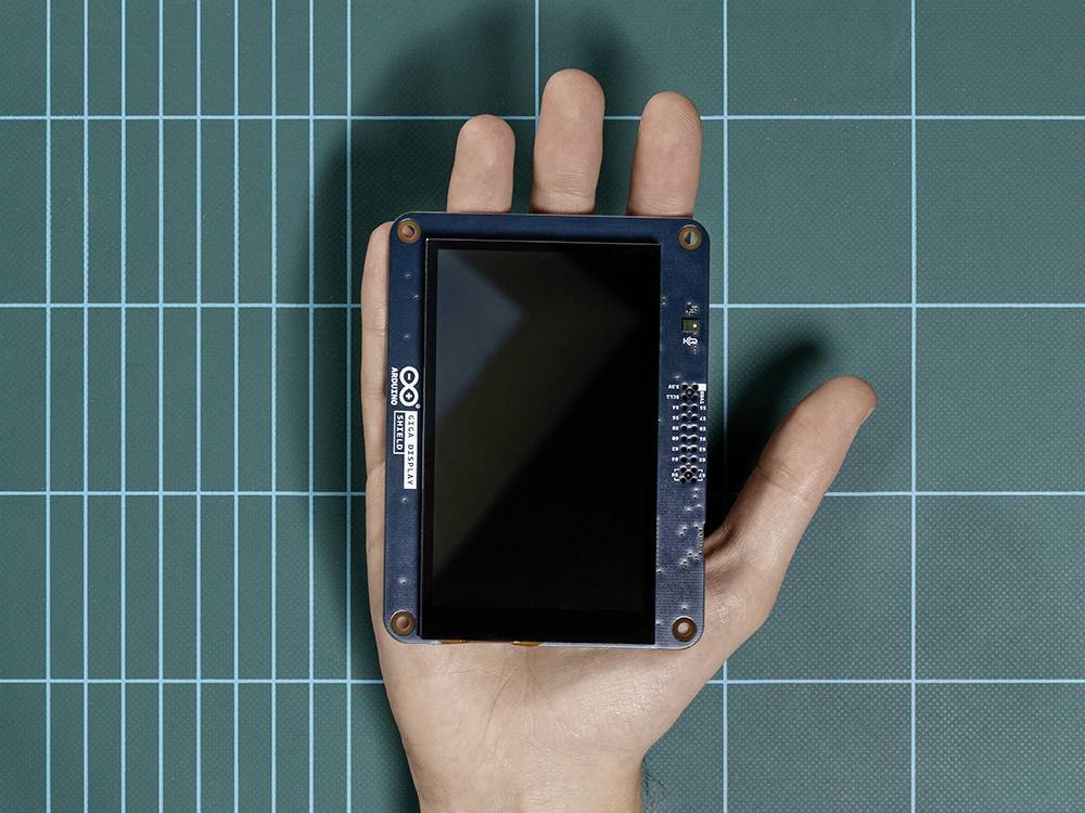 A hand is holding a small, rectangular electronic display against a green checked background.