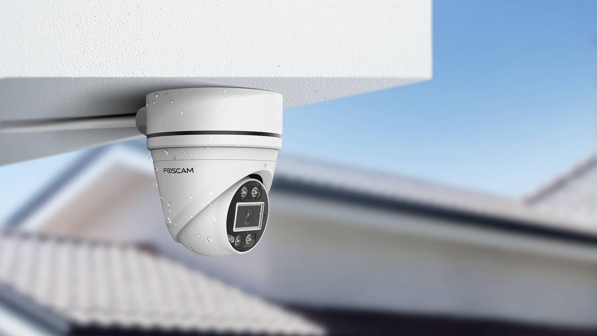 A white security camera is mounted on the exterior of a building. Blurred house roofs can be seen in the background.