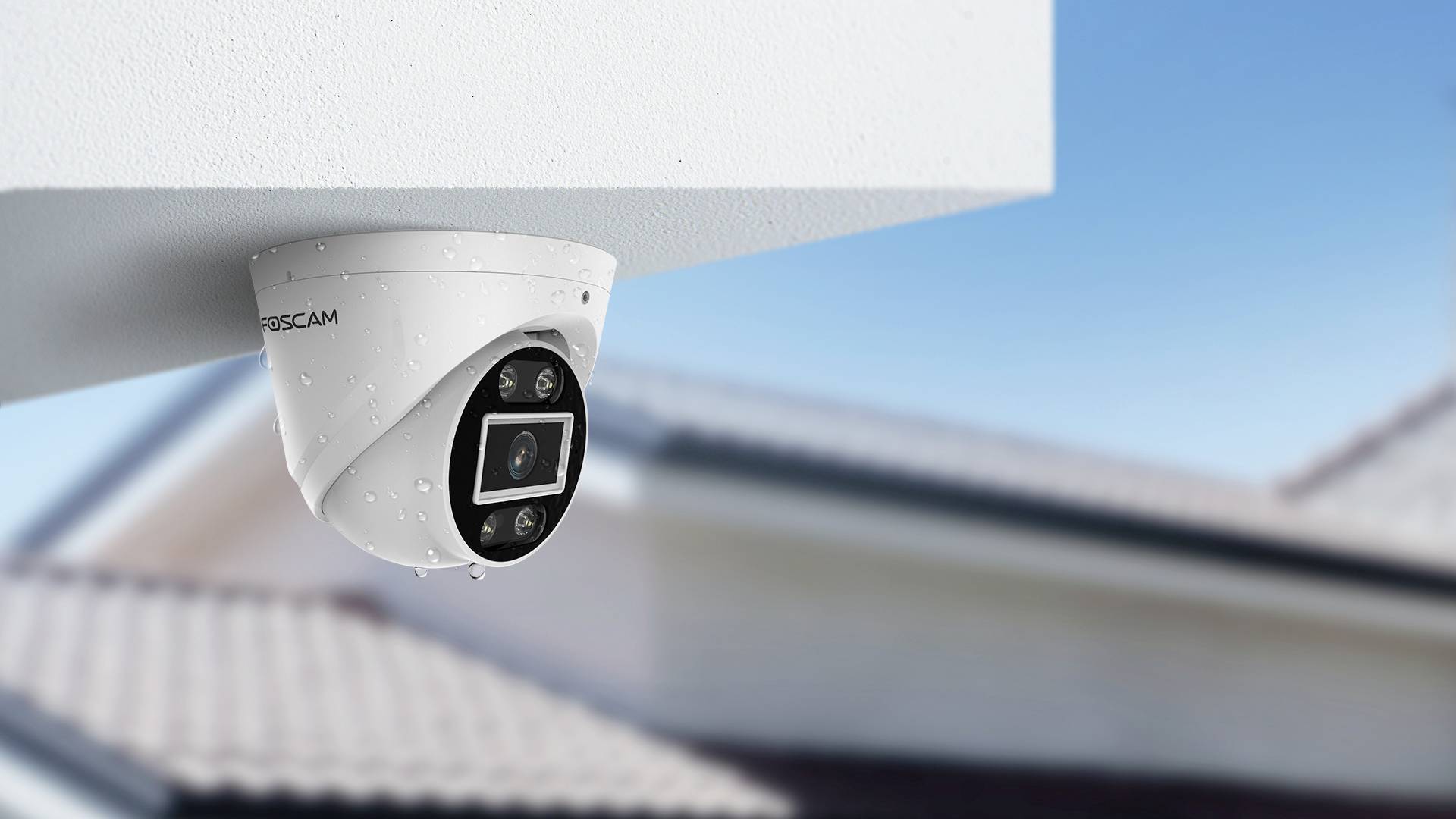 A security camera is mounted on the corner of a building, with a blurry roof in the background under a blue sky.