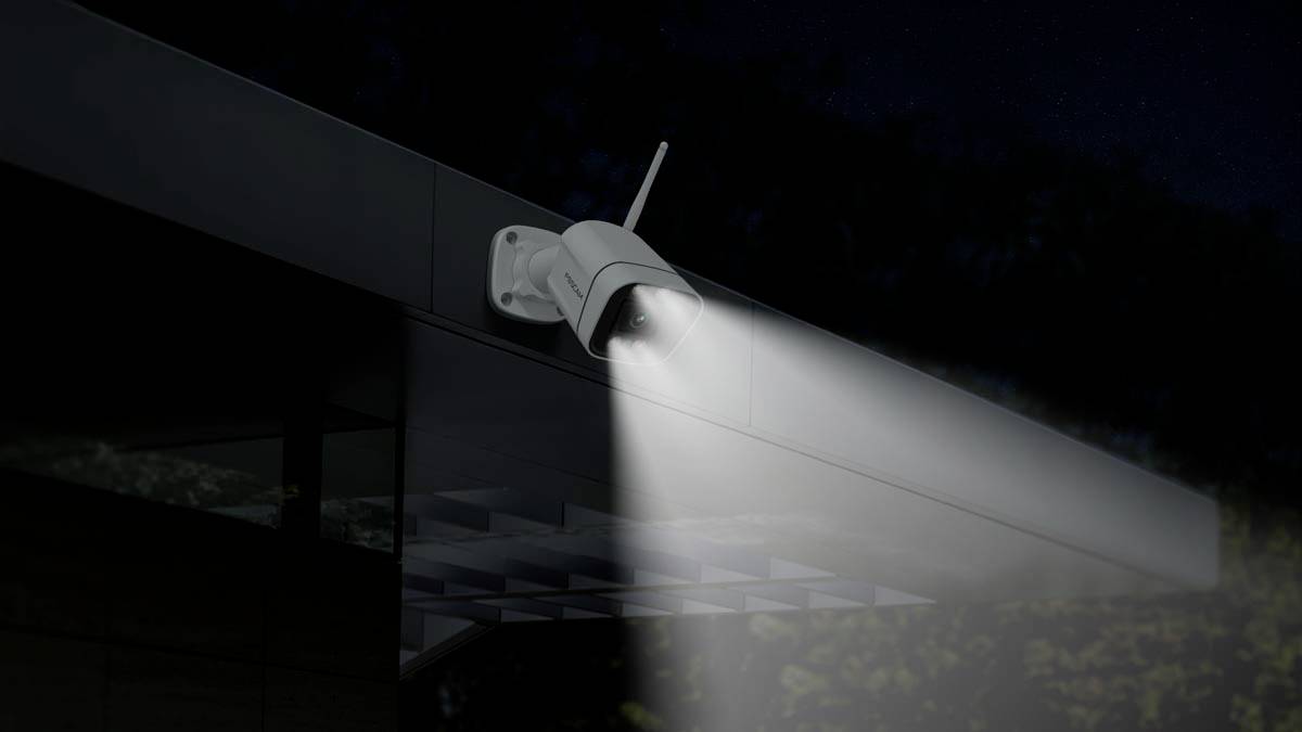 A security camera with an activated floodlight is mounted on the exterior of a building, illuminating the area beneath it.
