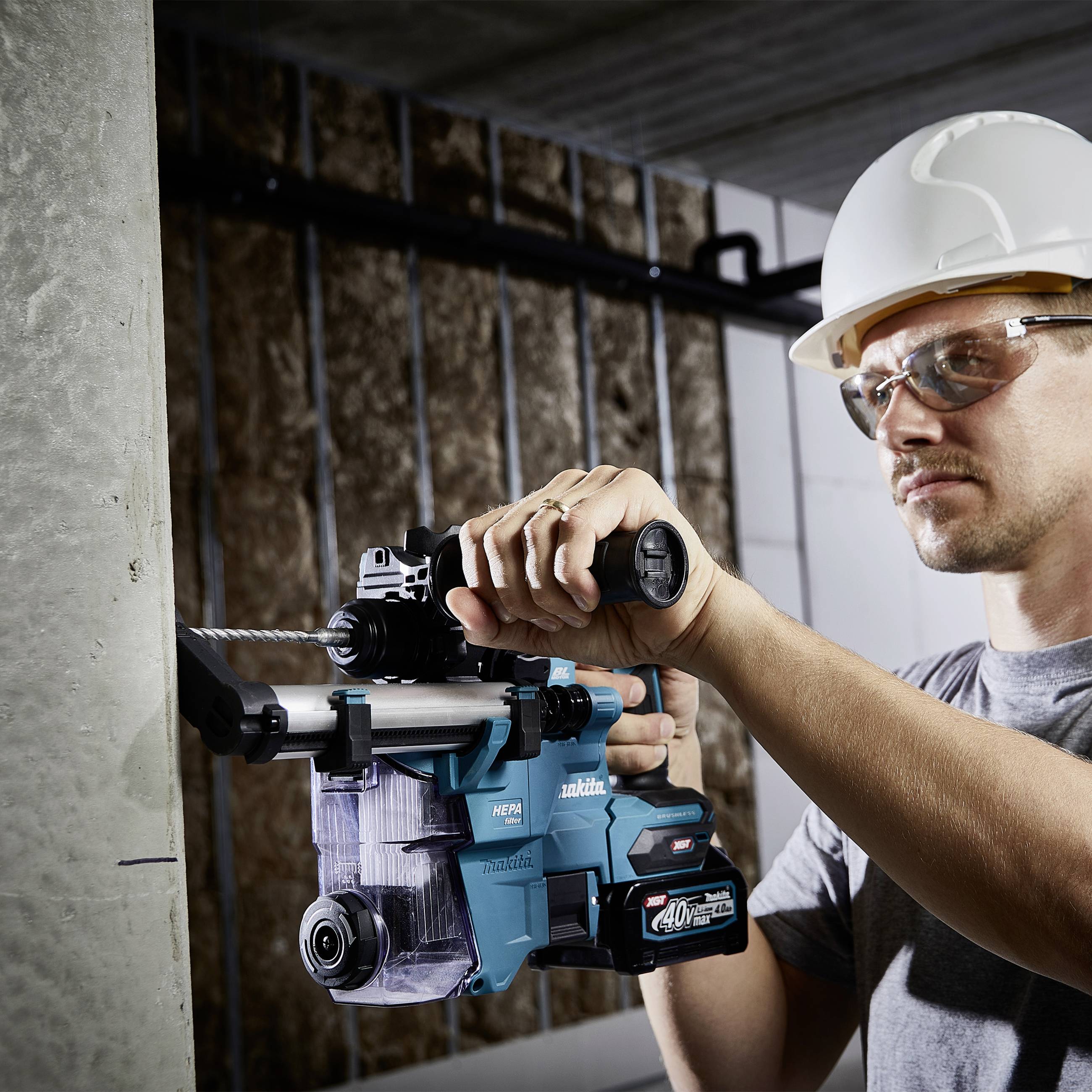A construction worker wearing a hard hat and safety glasses is using an electric tool to drill into a concrete wall.