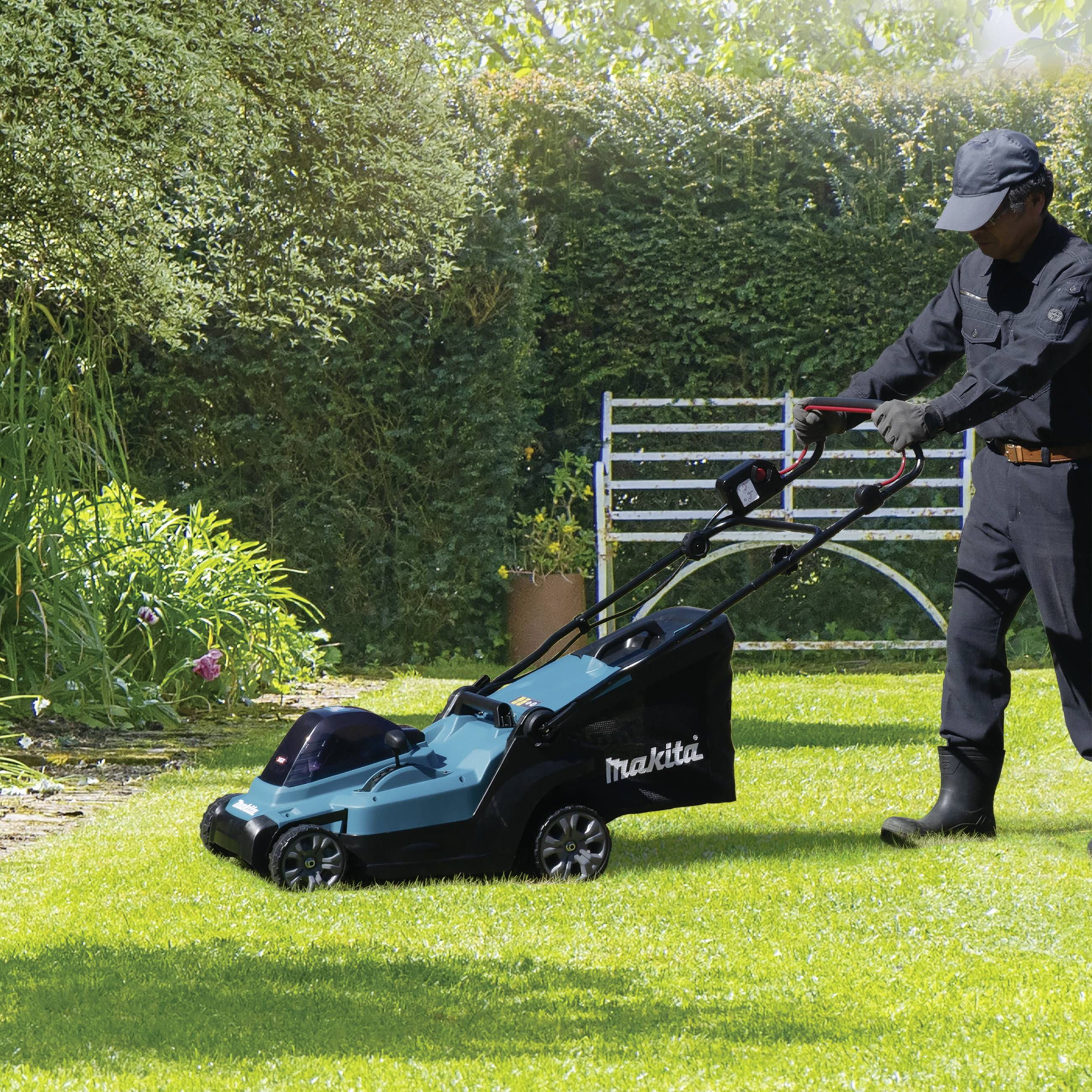 A person is mowing the lawn with an electric lawnmower on a sunny lawn; with a bench and green plants in the background.