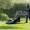 A person is mowing the lawn with an electric lawnmower on a sunny lawn; with a bench and green plants in the background.