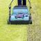 A person is mowing the grass with a blue lawnmower. On the left is tall grass, on the right is an already mown strip.