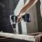 A hand is holding a blue cordless drill and drilling a hole into a wooden board. Wood shavings fall downwards as the drill operates.