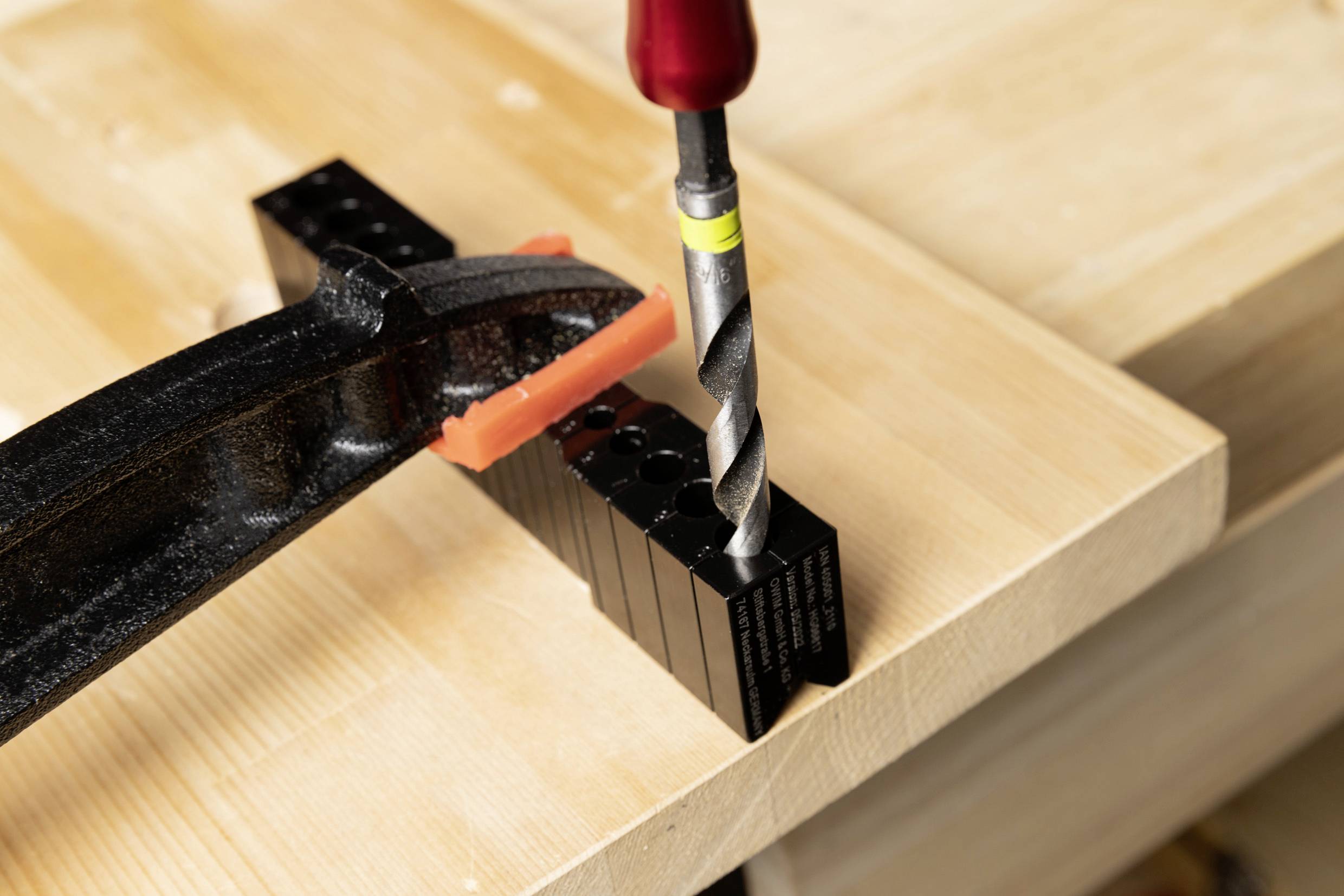A black plastic part is secured with a clamp on a wooden table. A drill is aligned above the plastic.