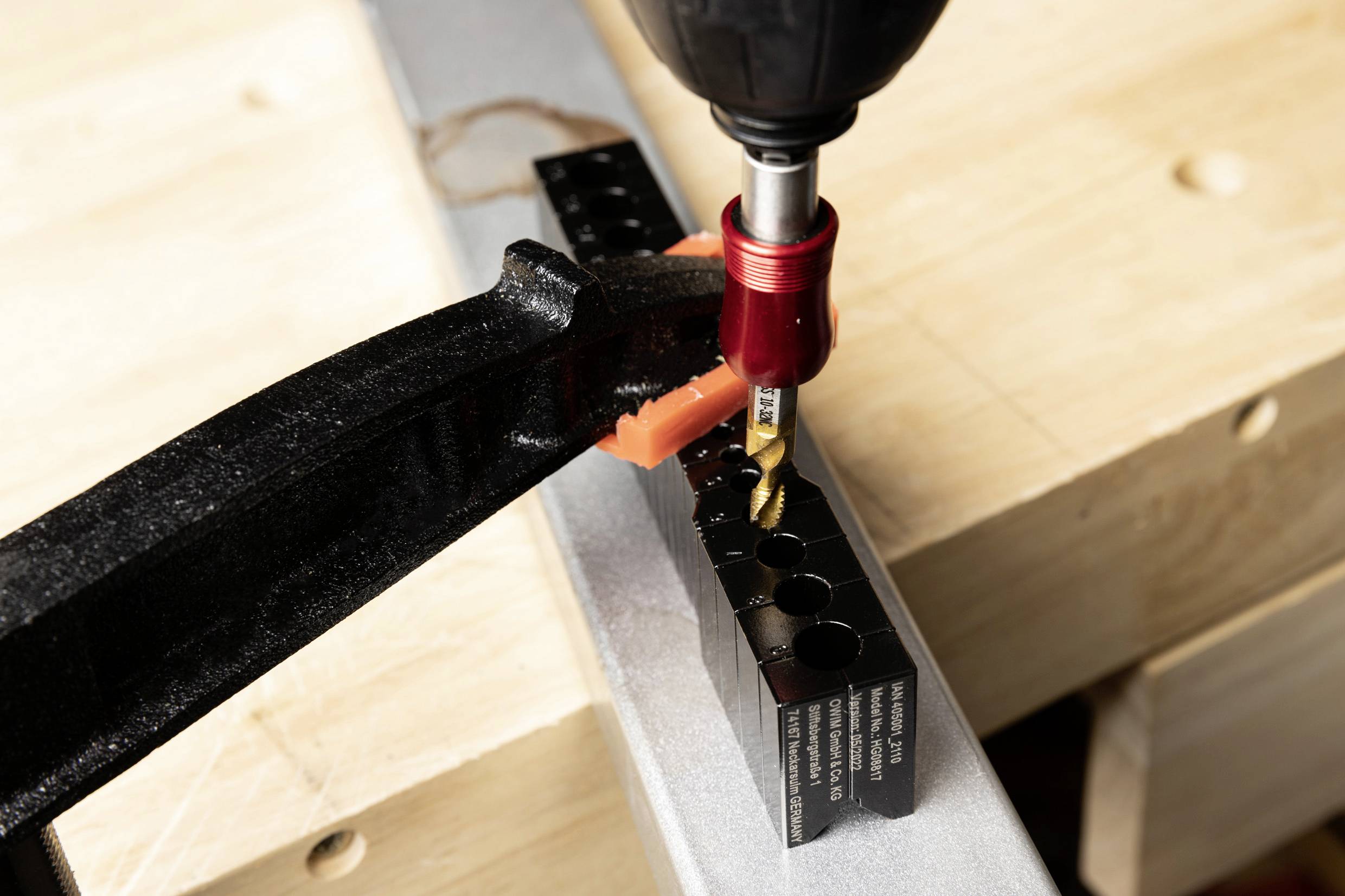 A drill with a metal drill bit is drilling a hole into a black metal piece that is secured with a clamp on a wooden table.