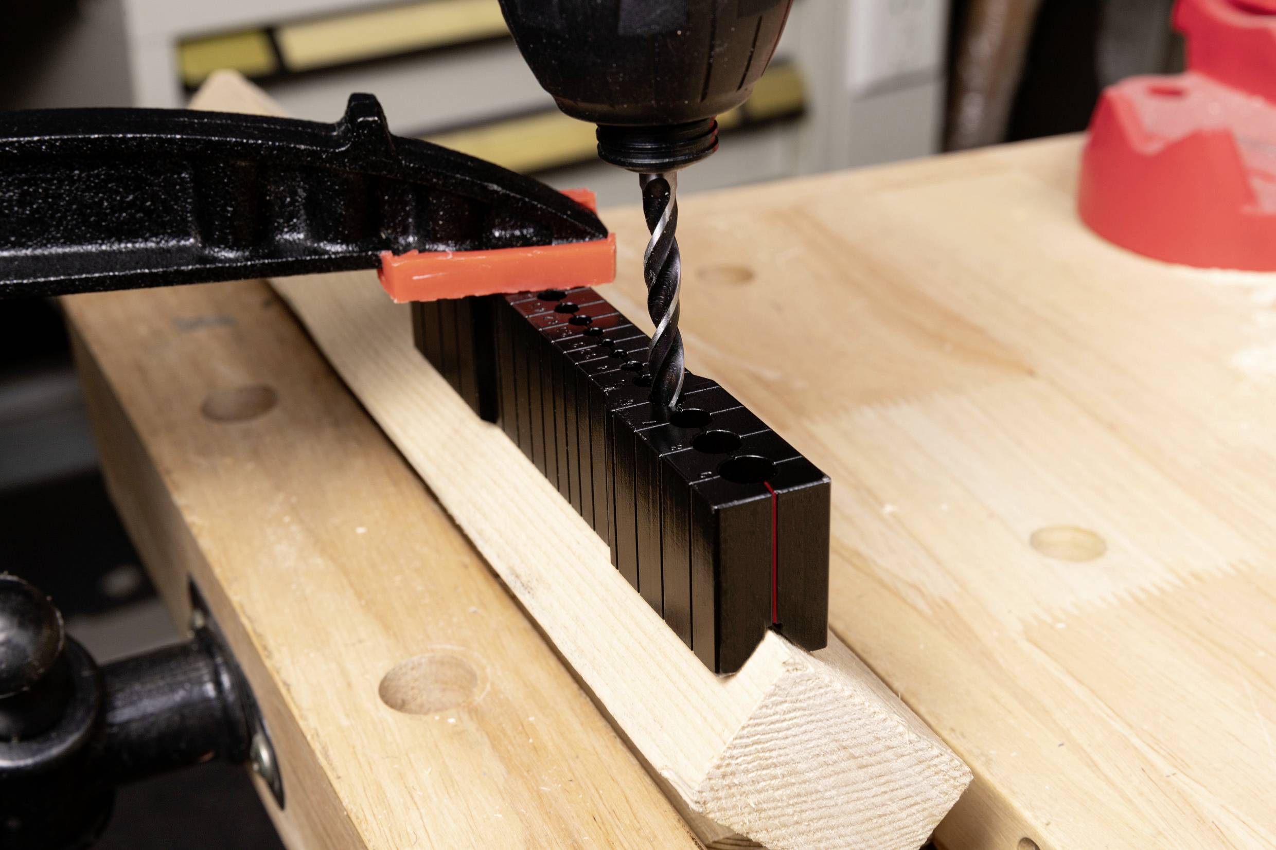 A drill is drilling holes into a wooden beam, secured by a black clamp. Workplace in a workshop.