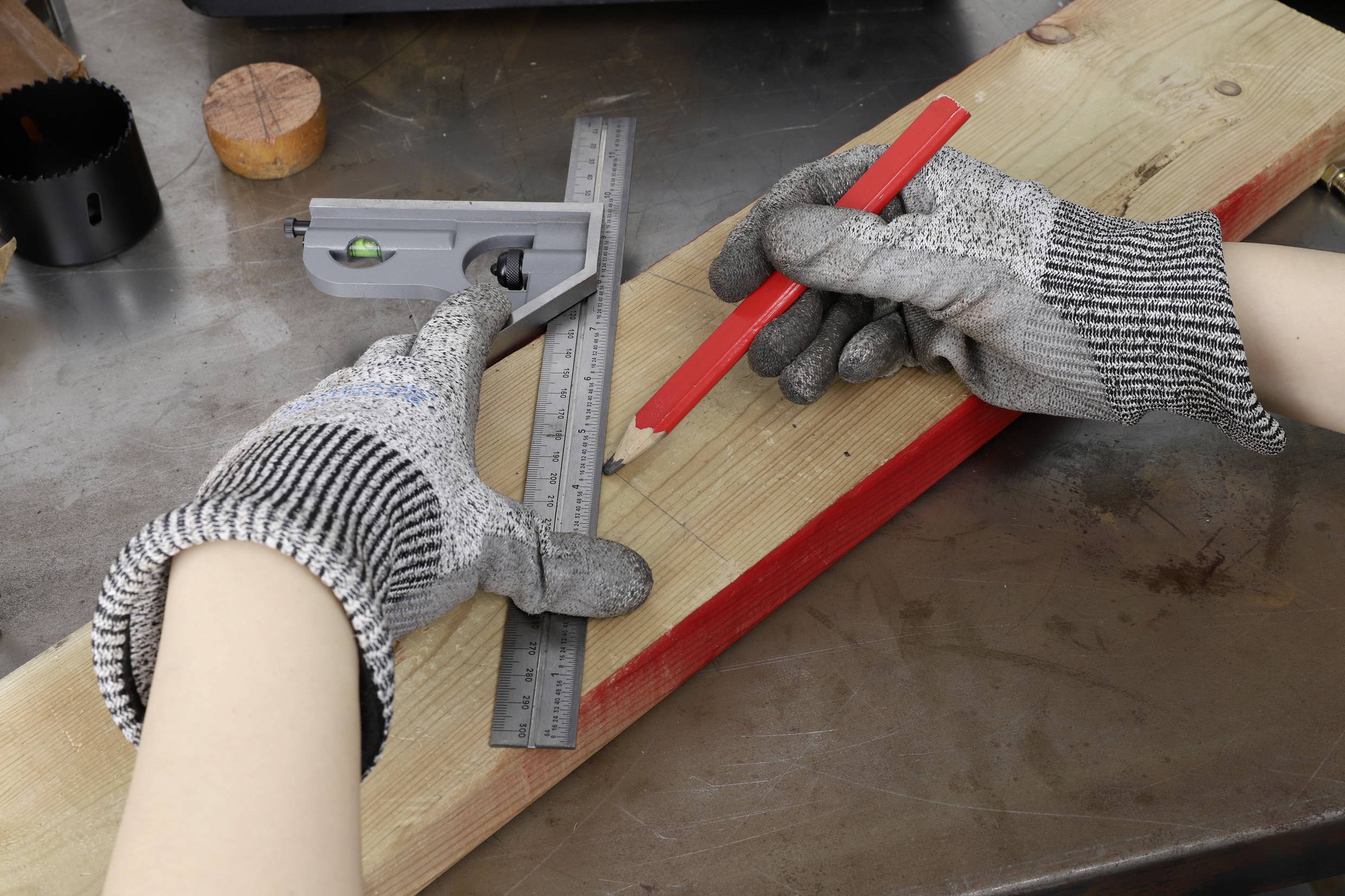 A person wearing gloves is marking a wooden board for cutting using an angle and a red pencil.