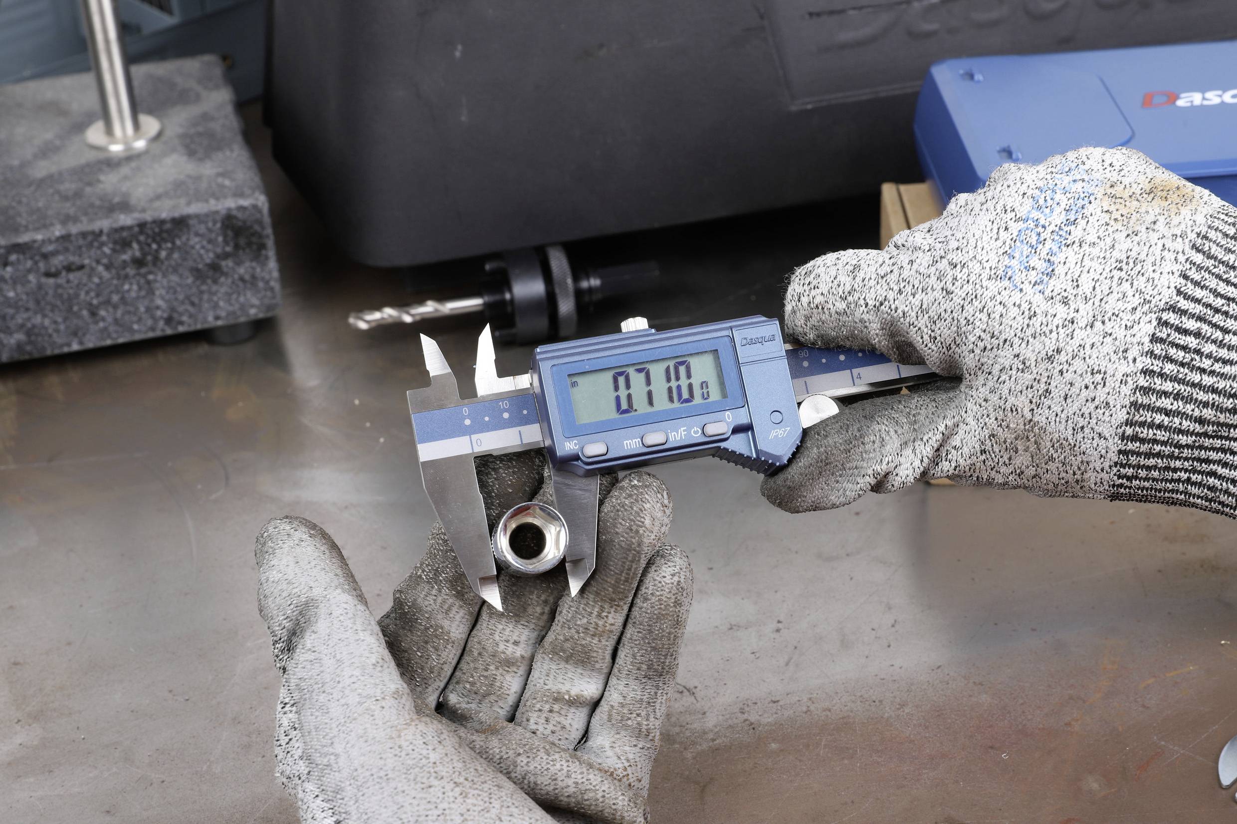 A person wearing gloves is measuring a metal part with a digital calliper. The display shows '7.10'. The background shows workshop supplies.
