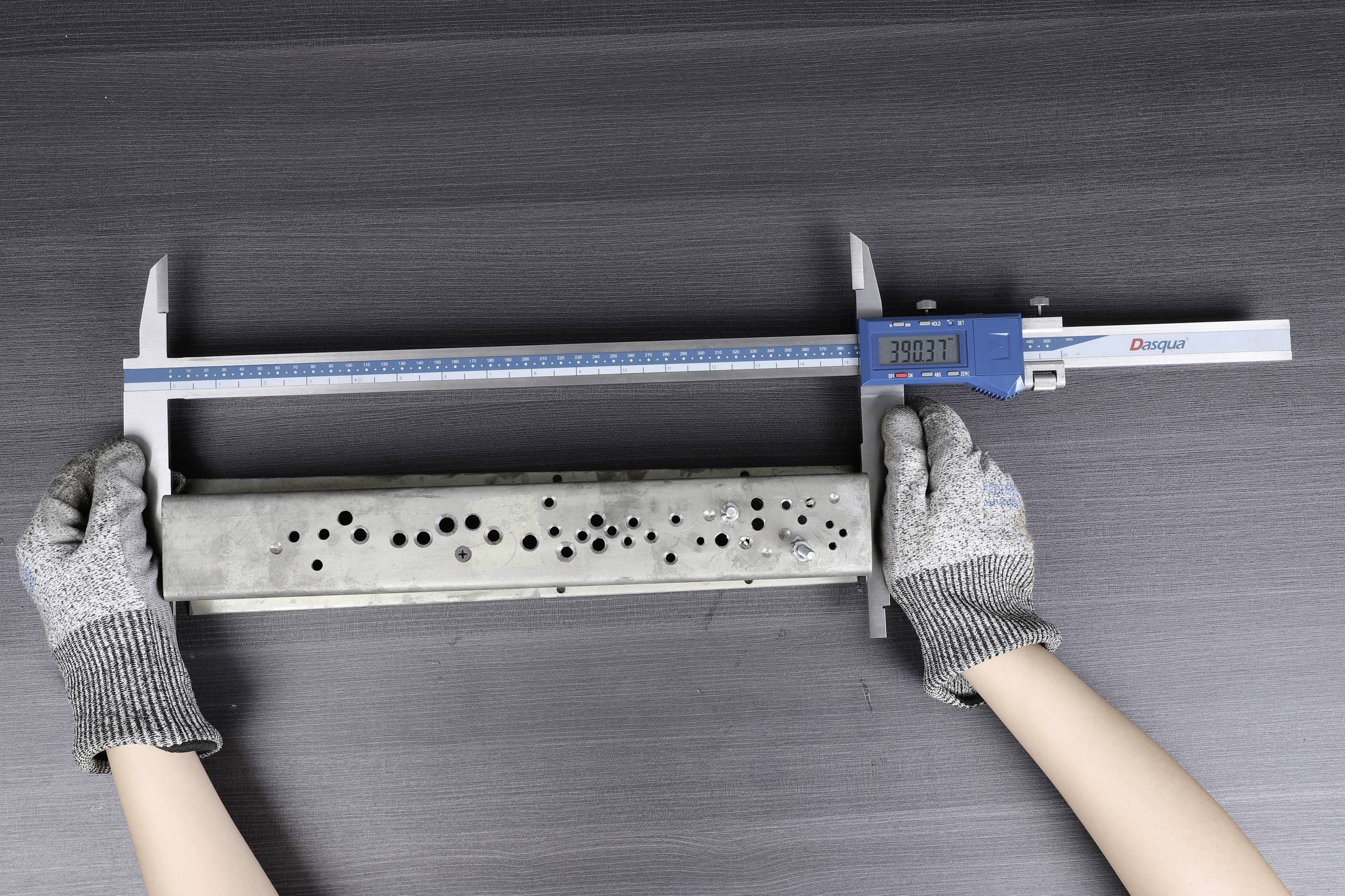 A person wearing grey gloves is measuring a perforated metal piece with a digital calliper against a dark background.