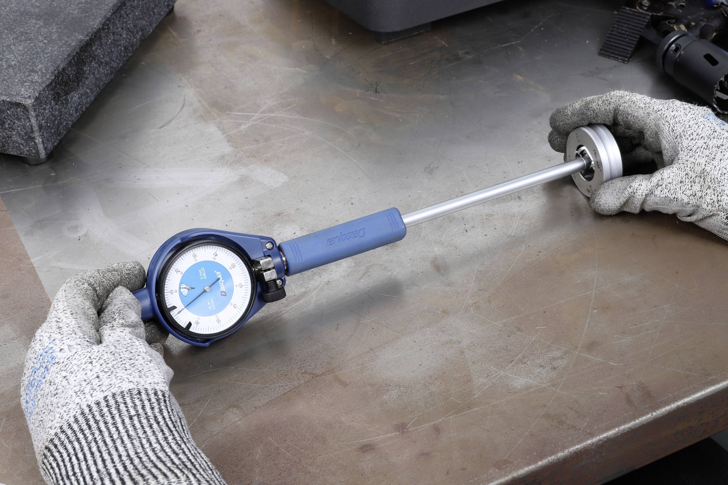 Two hands wearing gloves are holding a tool to measure the internal diameter of cylindrical objects on a metal table.