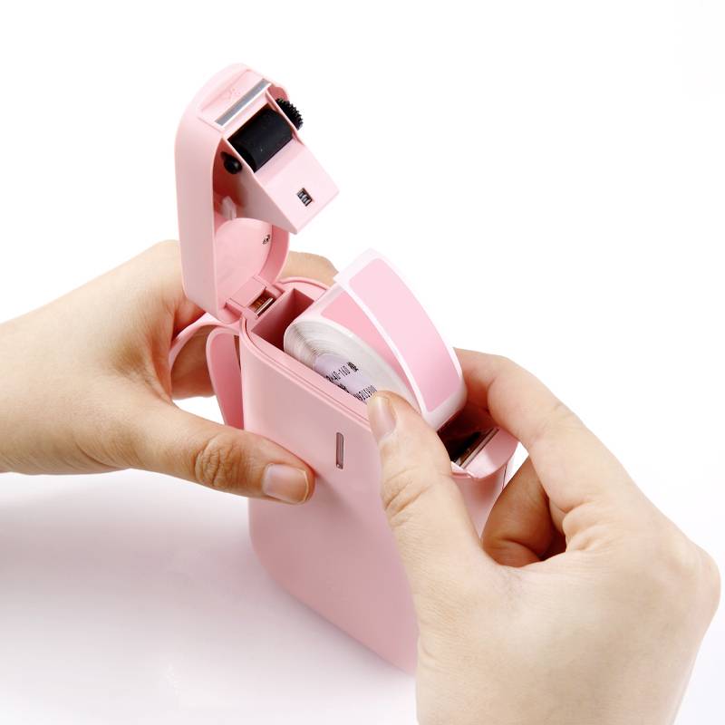 A hand is holding a small, pink label printer that is open. A user is changing the labelling roll in the device.