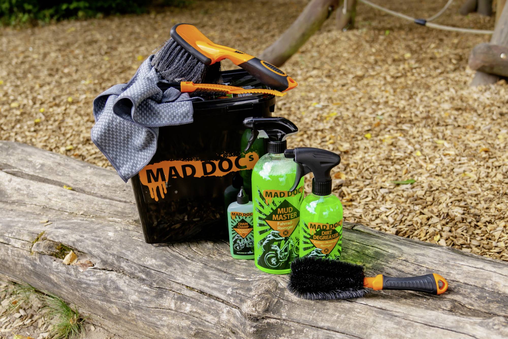 Bucket of cleaning products and brushes from 'Mad Doc's', placed on a wooden log, surrounded by wood chippings.