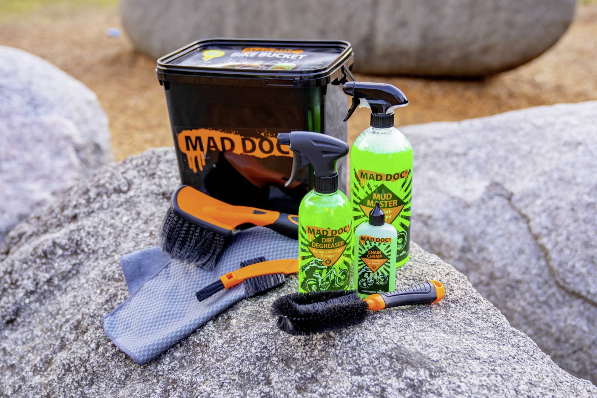 Cleaning set on a stone surface: a bucket with a 'Mad Doc' logo, two brushes, a cloth, and two bottles of cleaning solution.