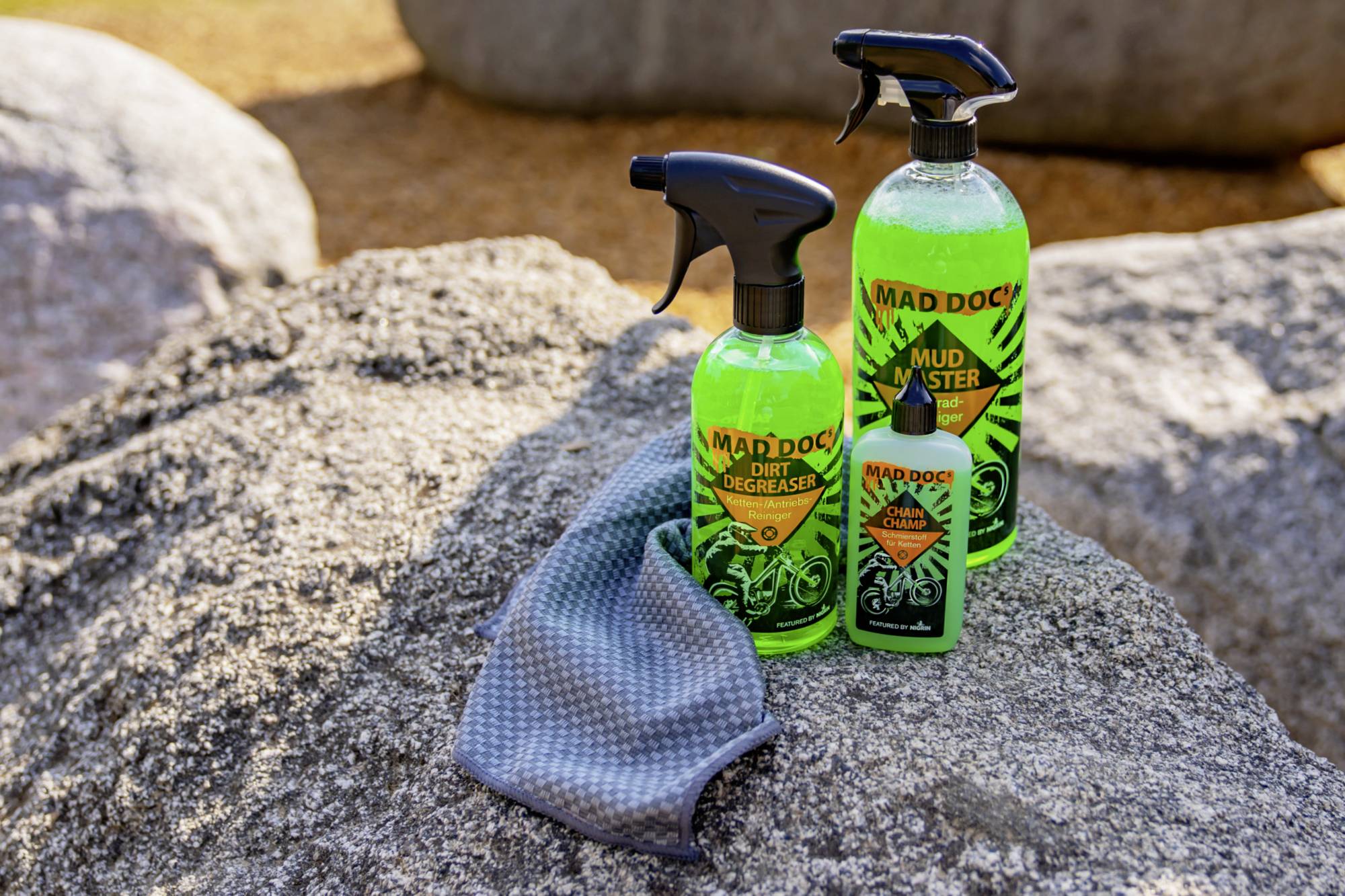'Three bottles of MAD DOG cleaning products and a cleaning cloth on a rock. Included are Dirt Degreaser and Mud Buster.'