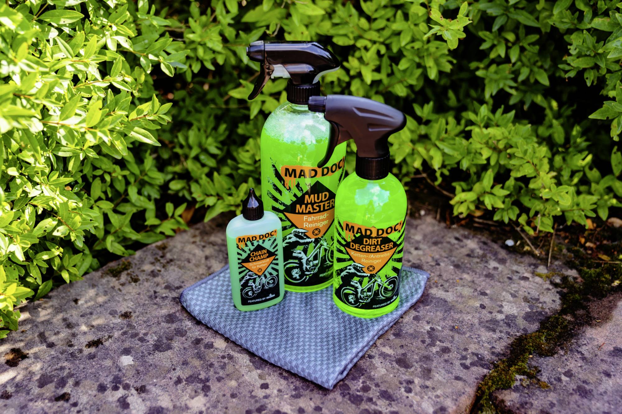 Three cleaning products in green spray bottles on a grey cloth, surrounded by green plants.