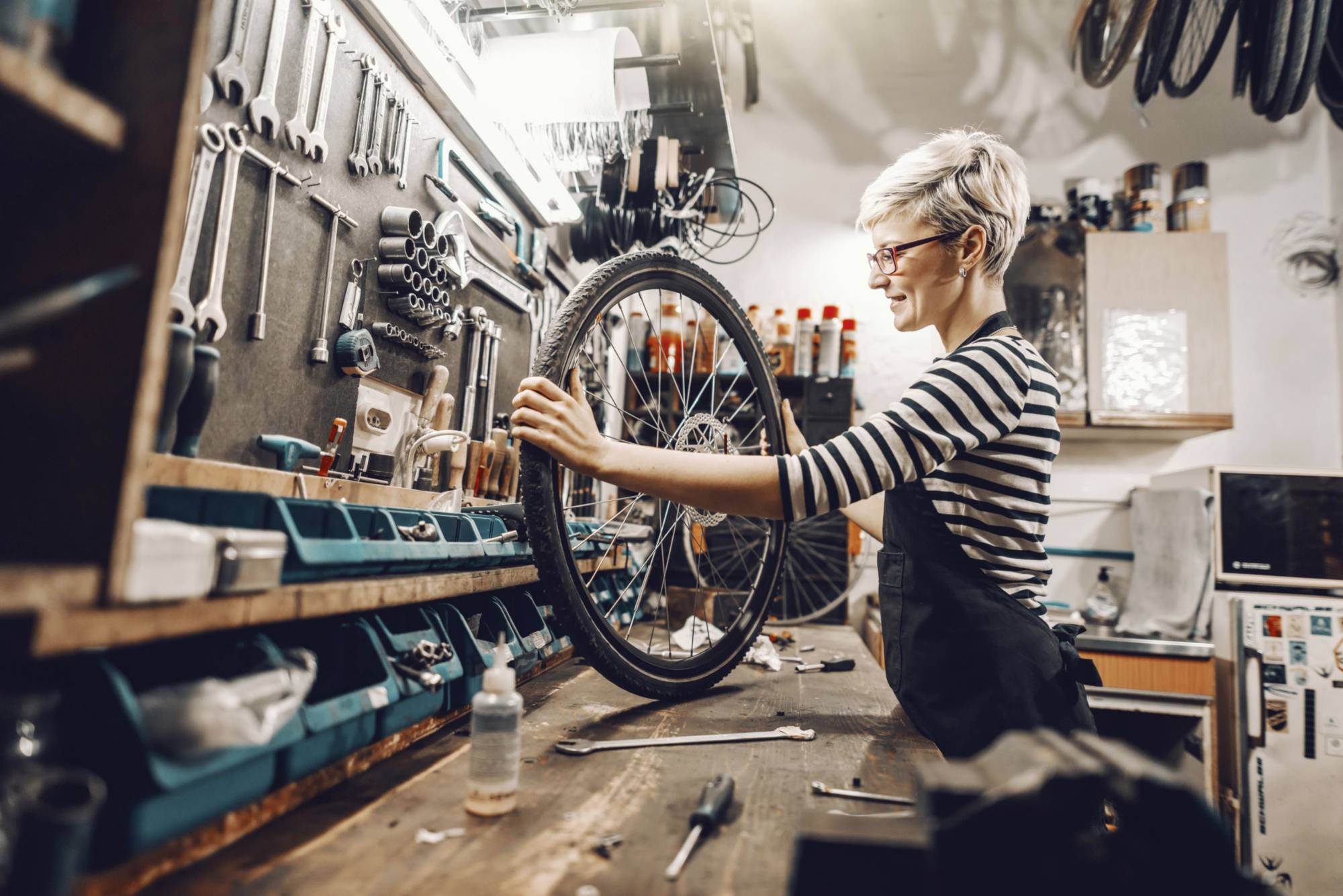 A person is repairing a bicycle in a workshop. Tools and spare parts are visible in the background.