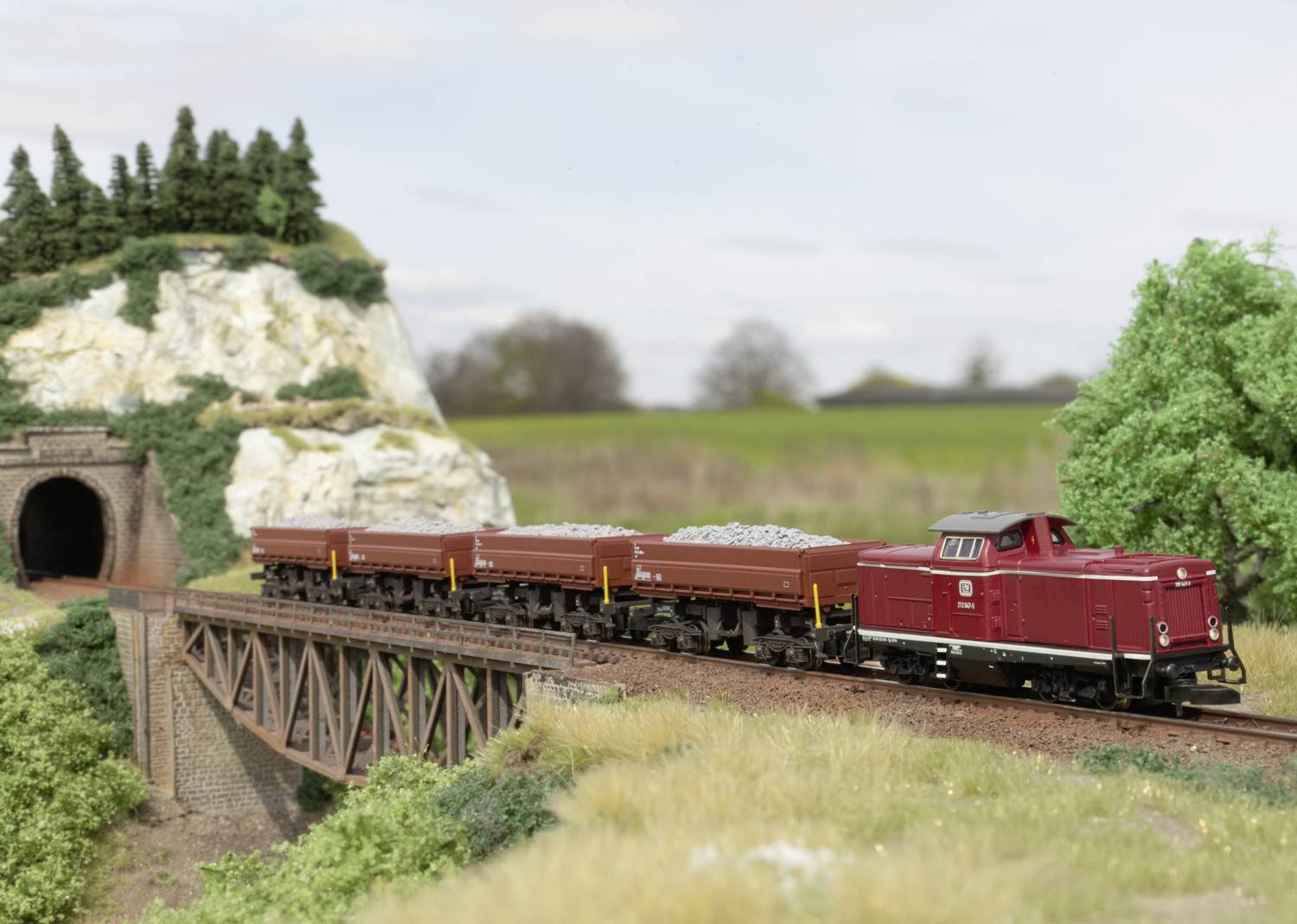 A red model train with open carriages travels across a bridge in a landscape with trees, a tunnel, and a hill.