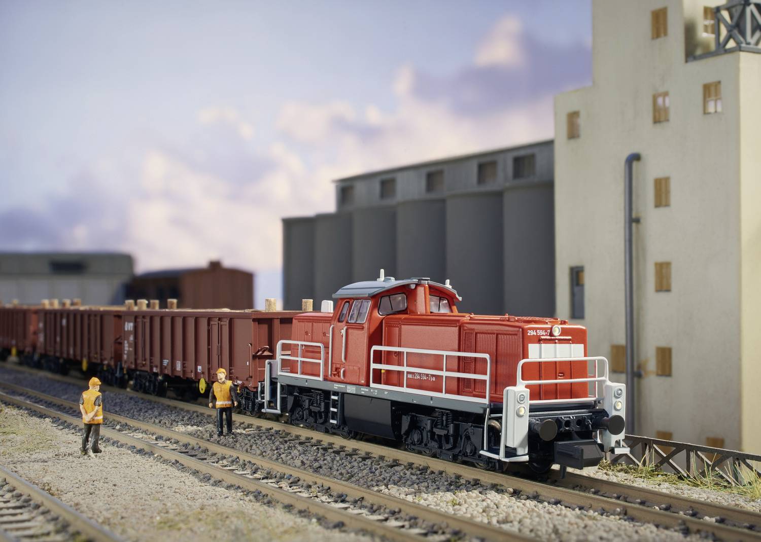 A red freight train on a track with two people in high-visibility clothing standing beside it, in front of an industrial site under a cloudy sky.