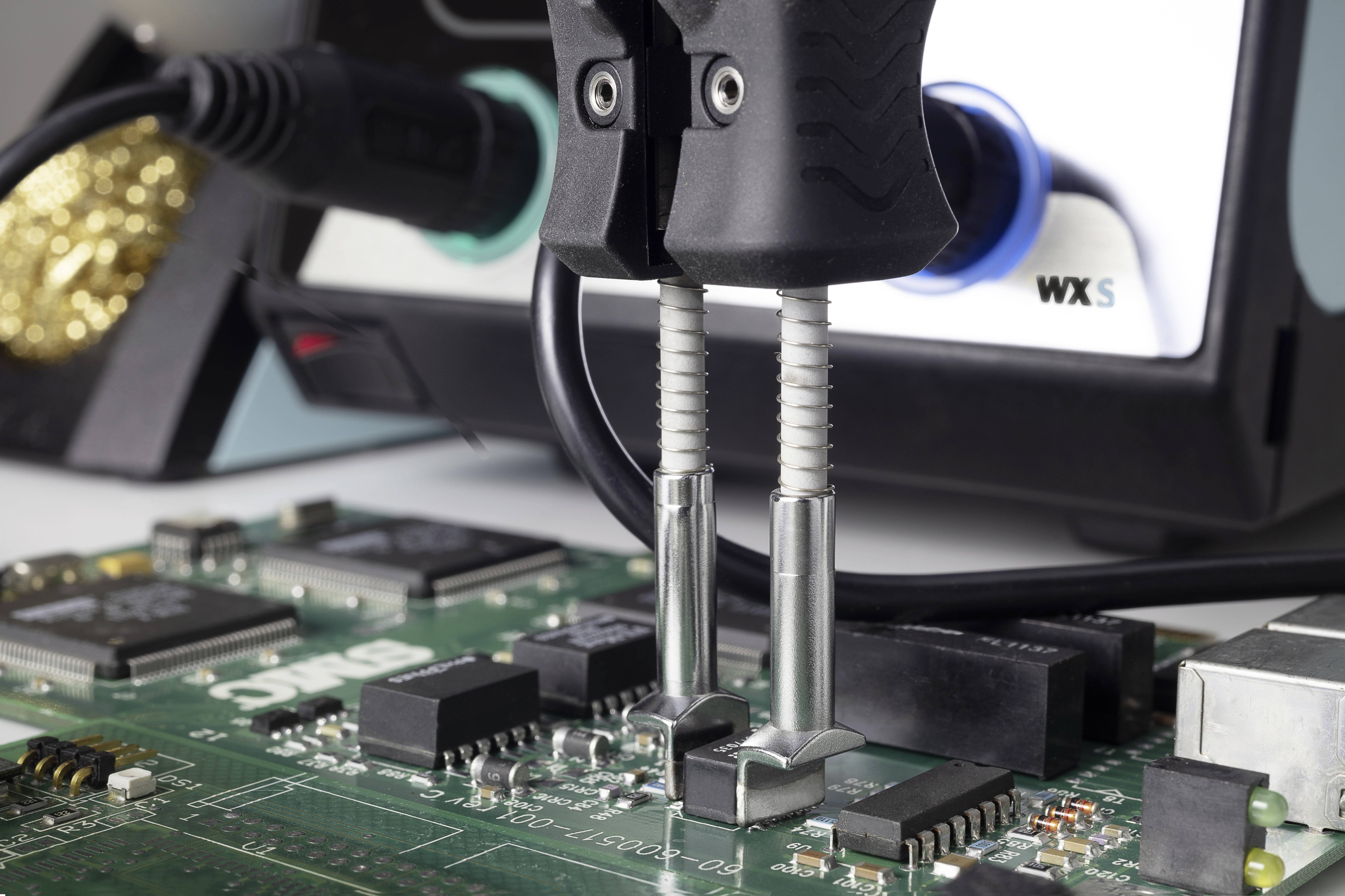 Soldering iron working on a printed circuit board. In the background, an electronic soldering station with a blue rotary knob is visible.