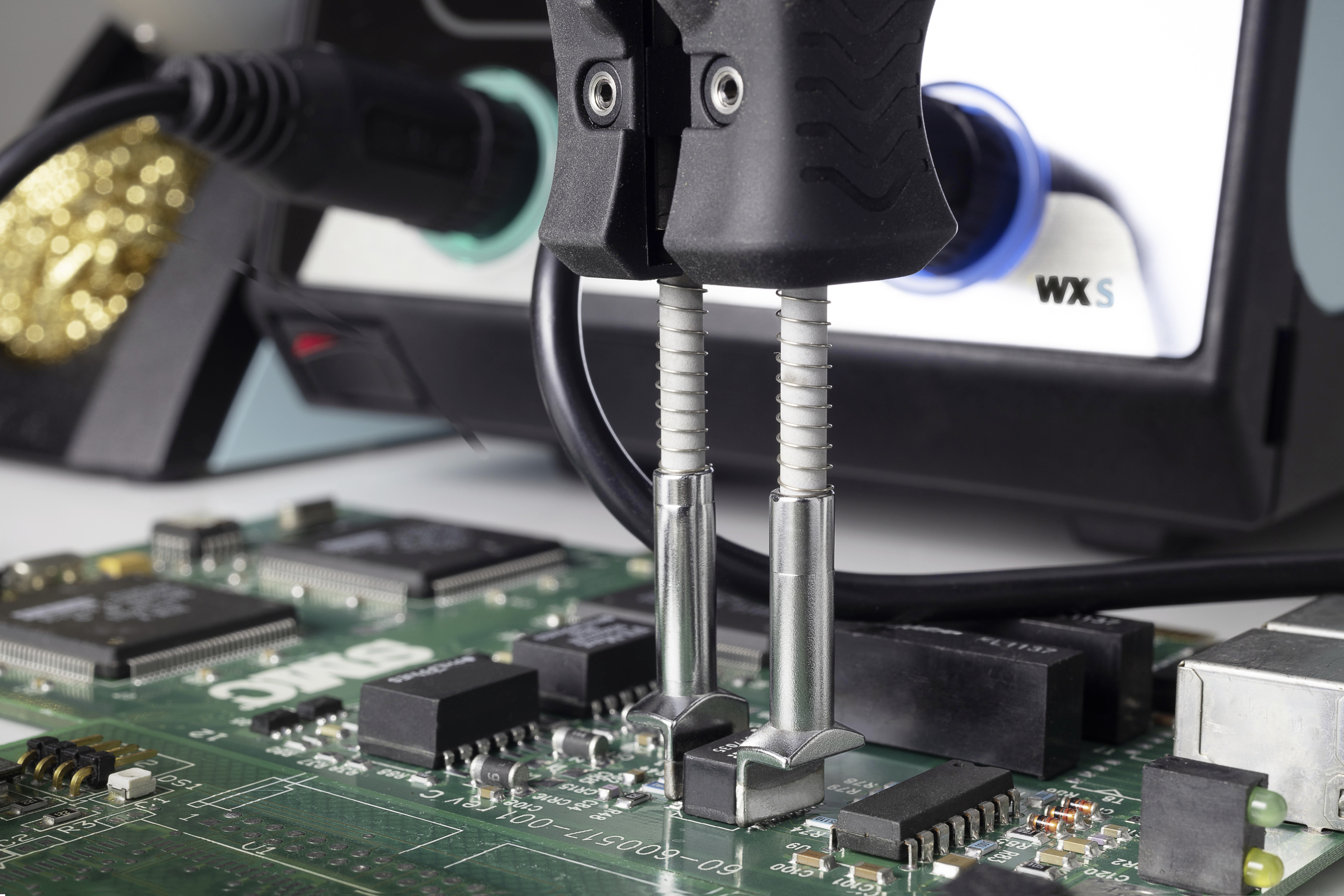Soldering iron working on a printed circuit board. In the background, an electronic soldering station with a blue rotary knob is visible.