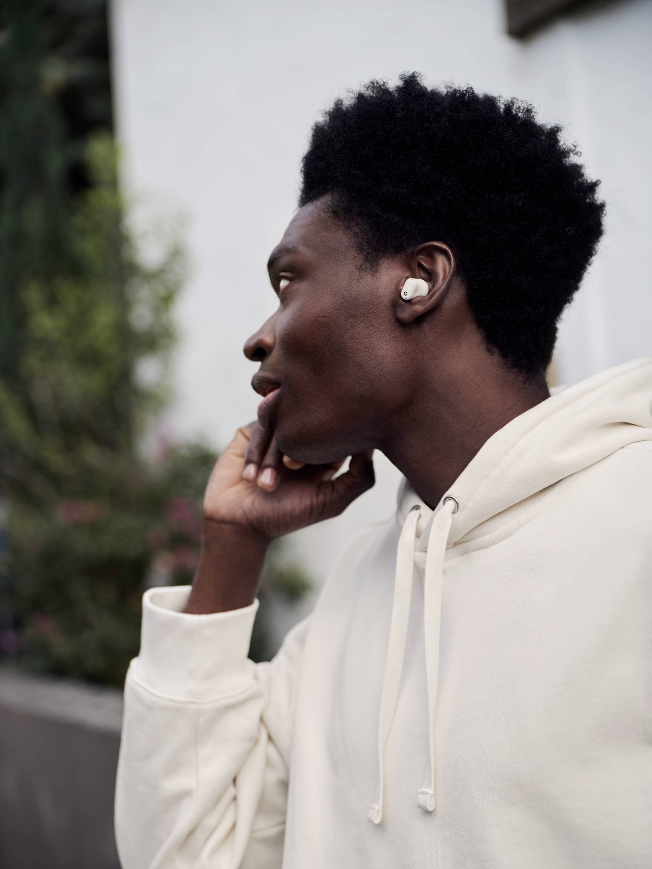 A person with a short haircut and white earbuds stands in profile, wearing a cream-coloured hoodie, with one hand resting on their face.