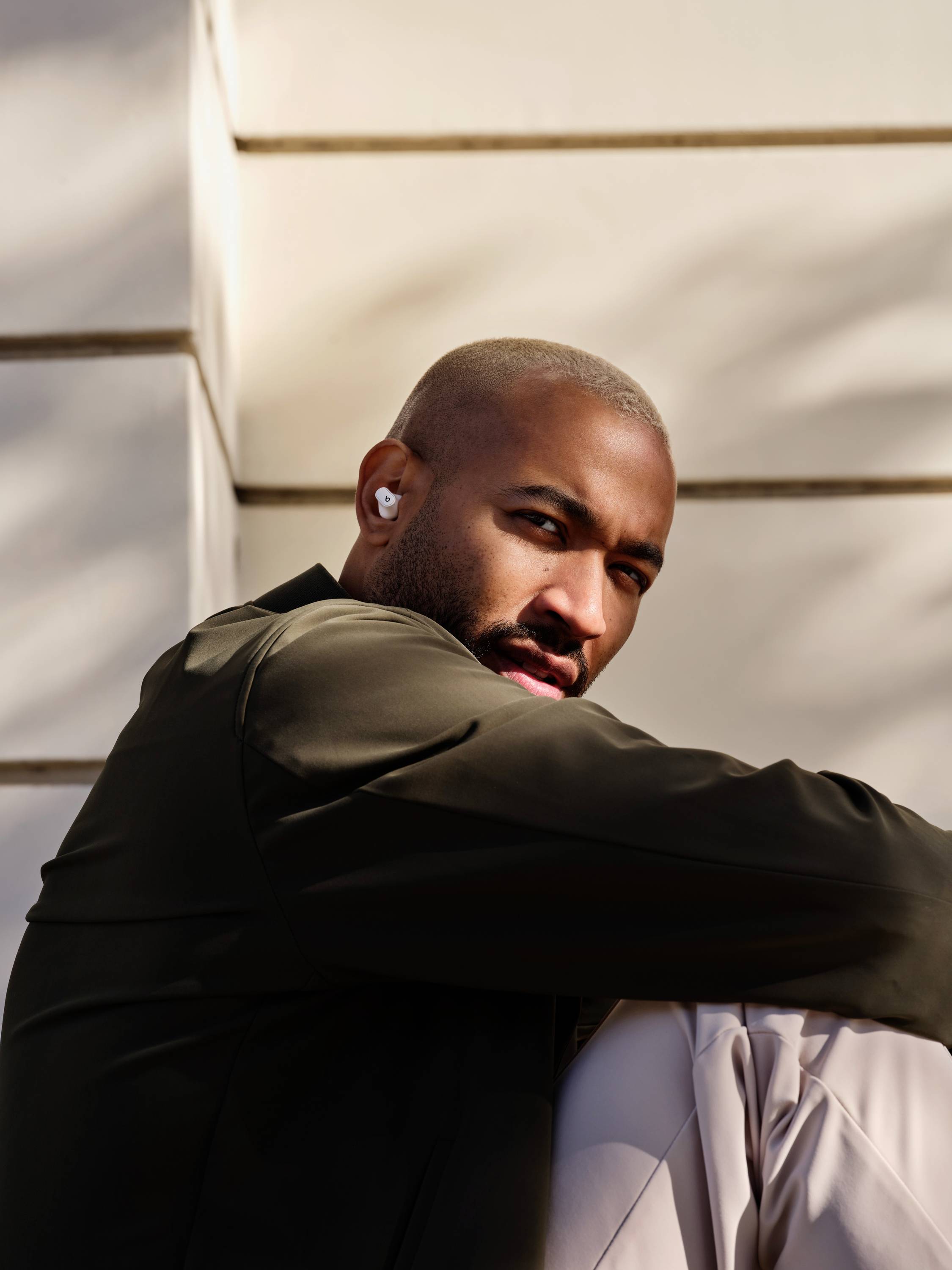 A person is sitting outside against a wall, wearing wireless earbuds. They are looking attentively to the side.