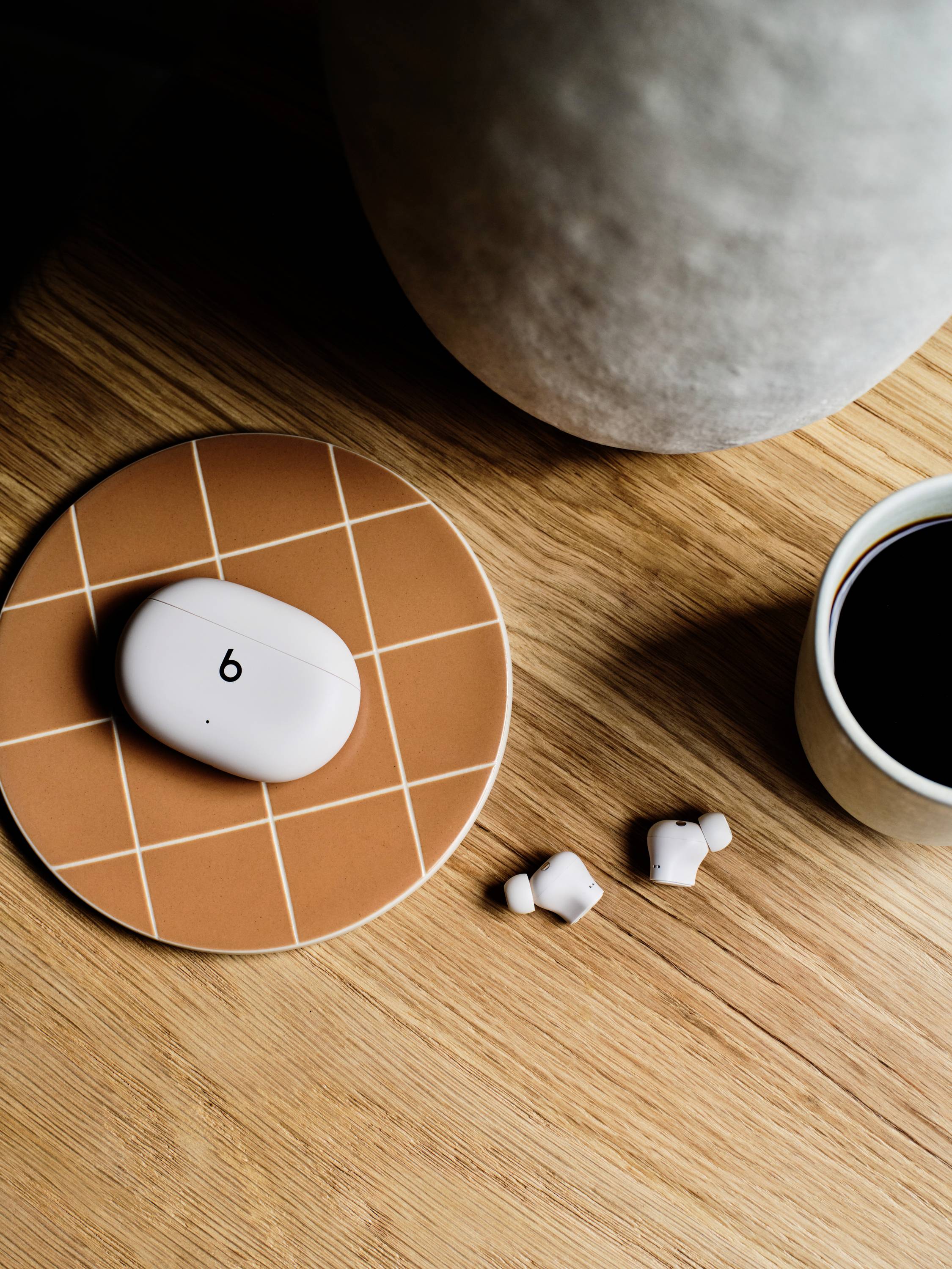 White wireless earbuds with charging case on an orange gingham coaster, beside a coffee cup on a wooden surface.