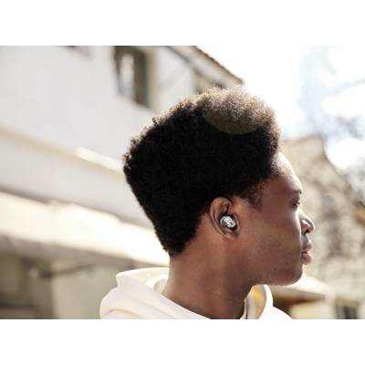 A person wearing earphones looks to the right, standing in a sunny outdoor area, with focus on the earphones.