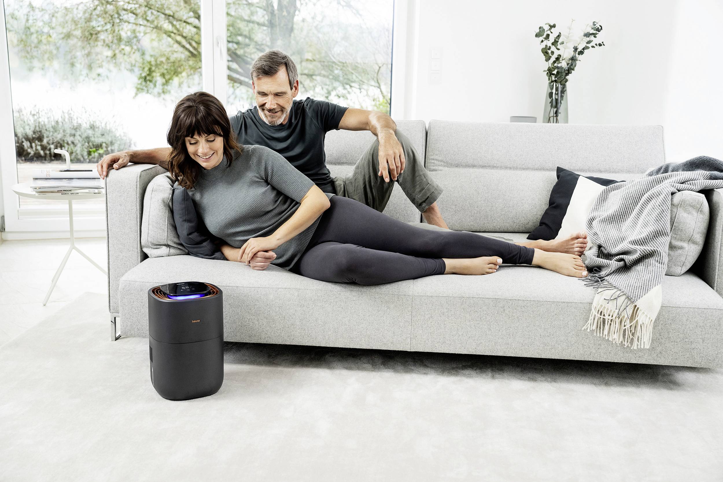 A woman is lying smiling on a sofa, a man is sitting beside her. An air purifier is standing in front of them. Outside, a green landscape is visible.