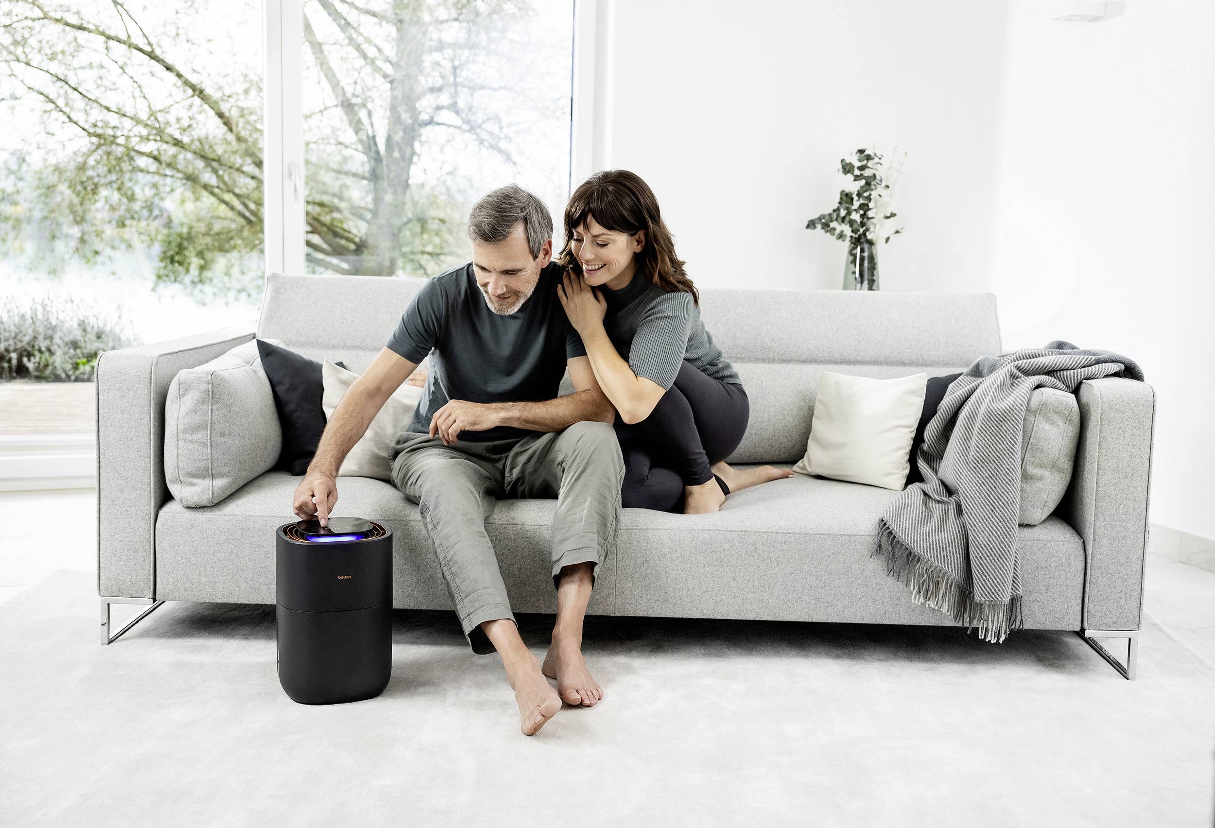 A couple sits on a grey sofa and looks at a small, black air purifier on the floor. In the background, a large window is visible.