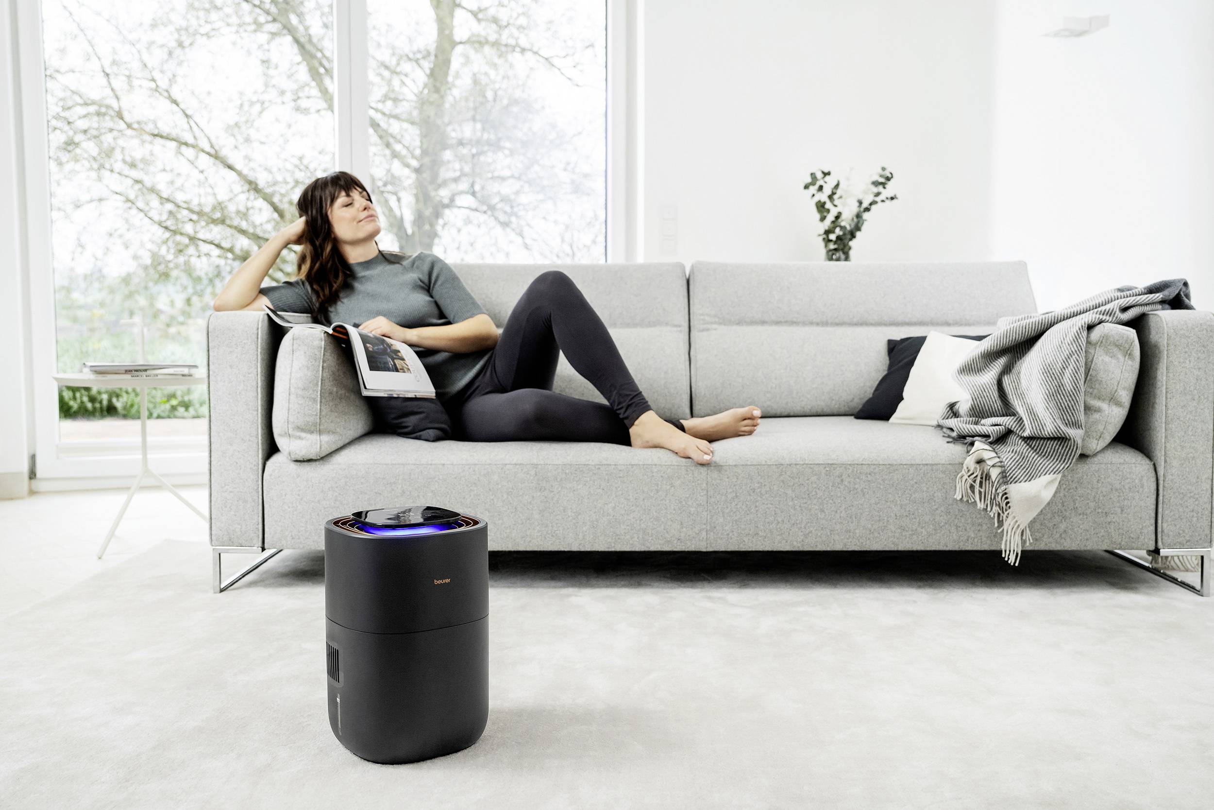A person is relaxing on a grey sofa, reading a magazine; a black air purifier stands in the foreground.